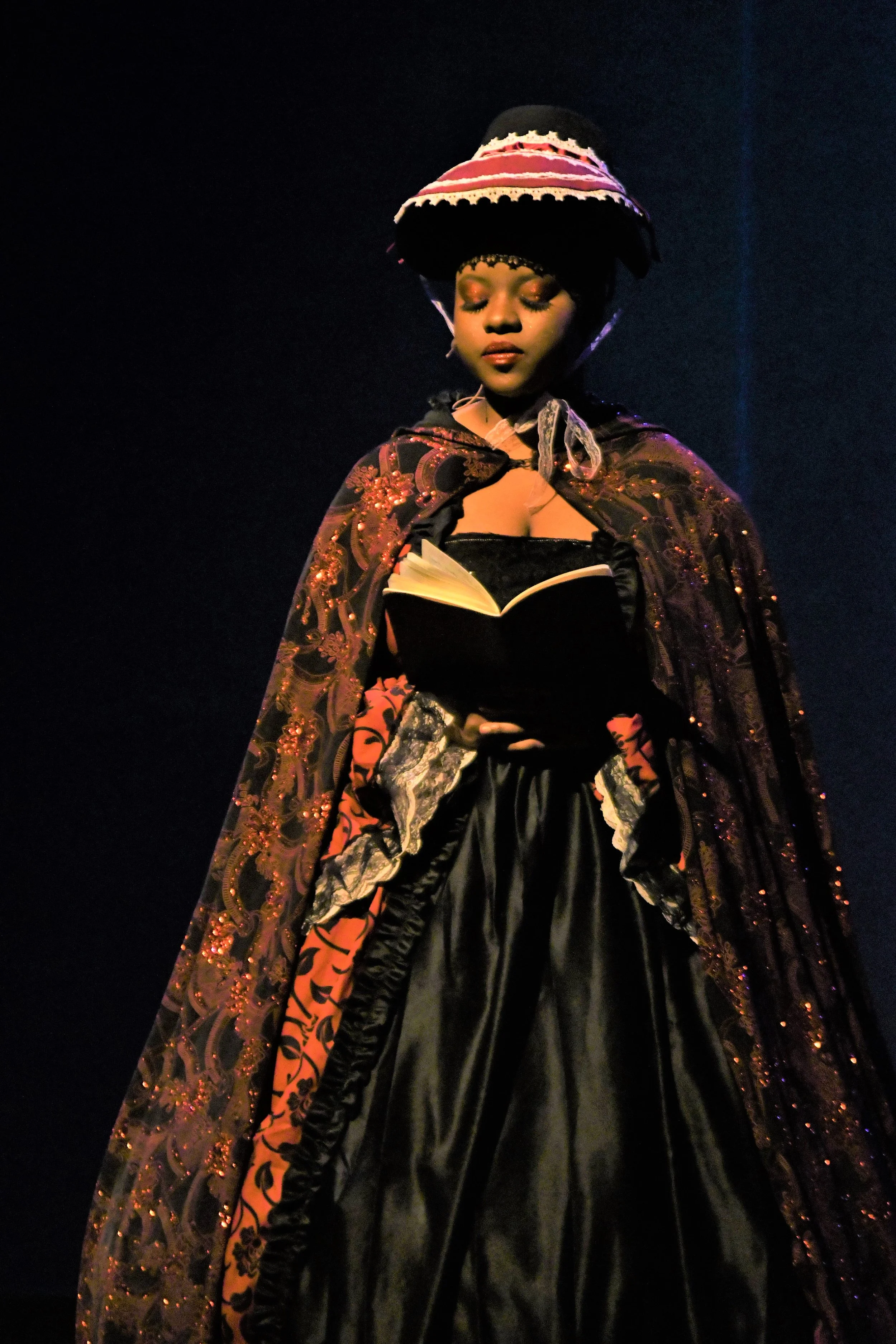 A woman dressed in historical costume holding an open book, wearing a large bonnet and a cape with intricate embroidery, standing against a dark background.