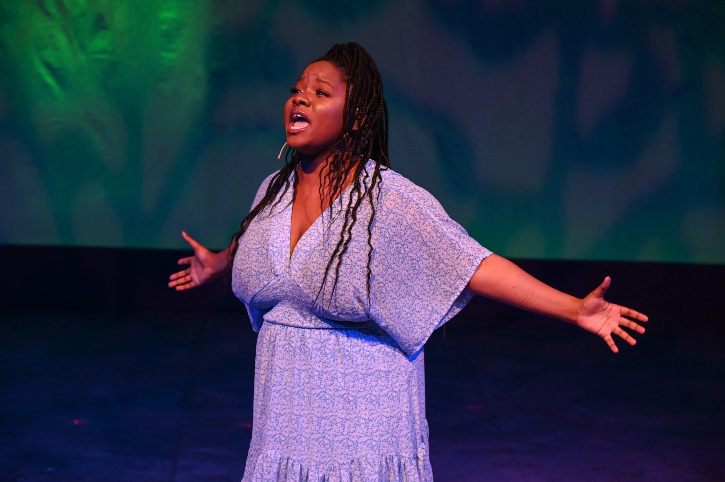 A woman with braided hair, wearing a light-colored dress with small floral patterns, appears to be singing or speaking passionately on stage with arms outstretched in front of a textured green and blue backdrop.