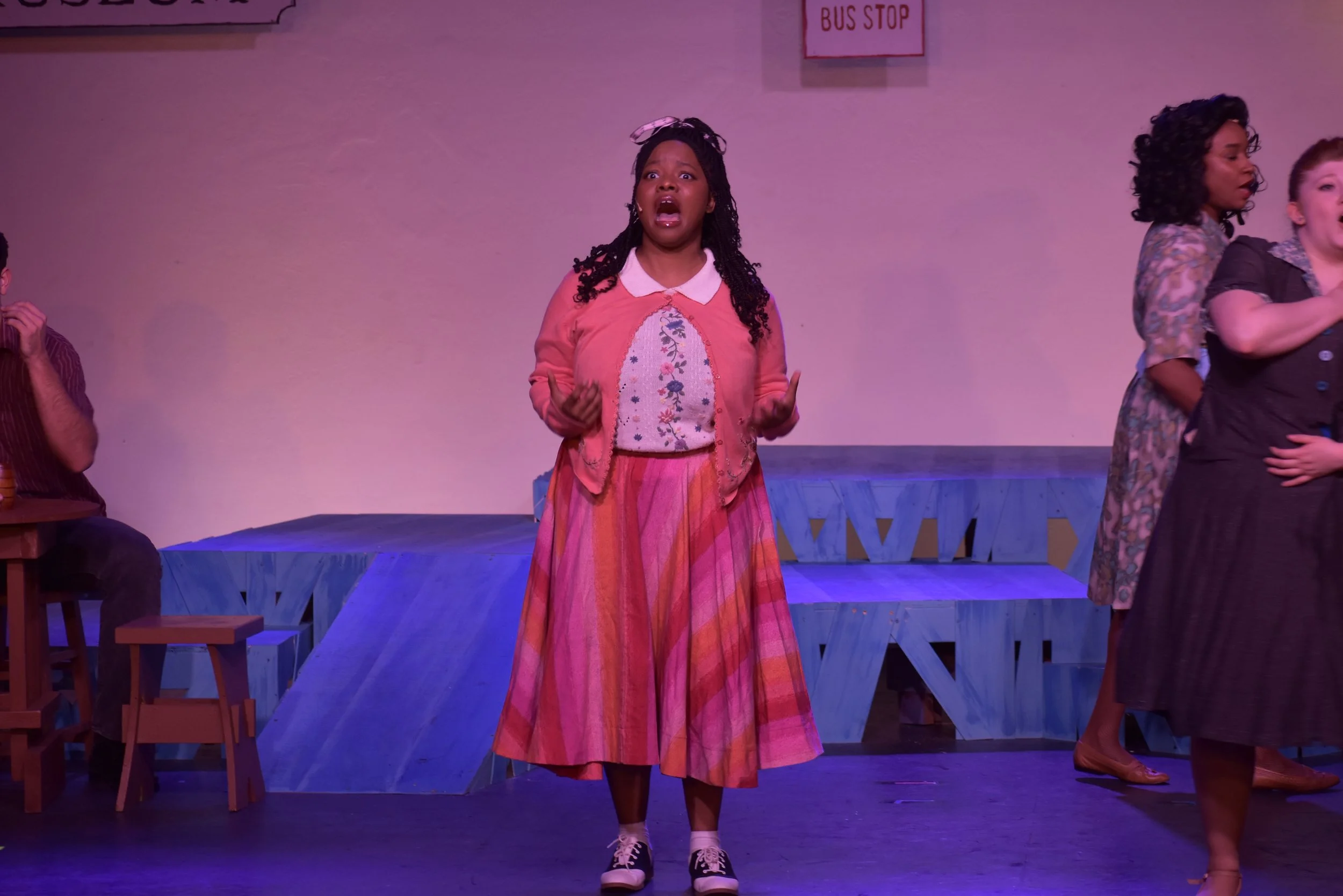 A young woman acting on stage with a surprised or emotional expression, wearing a pink cardigan, a floral blouse, a colorful striped skirt, and sneakers, during a theatrical performance.