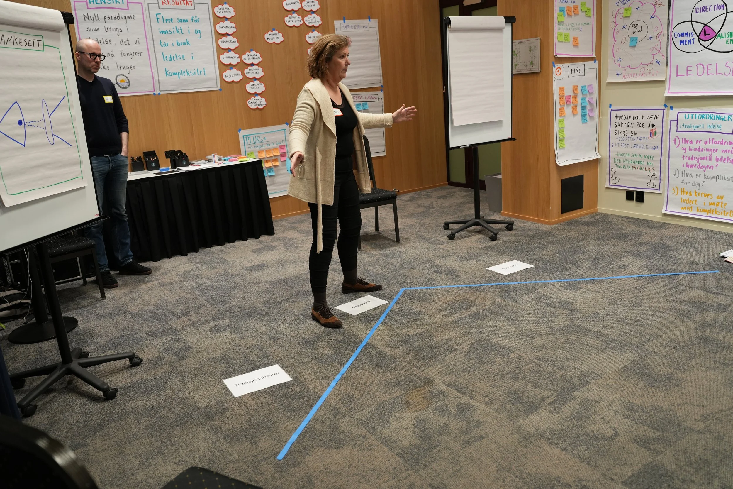 Kursholderne Anne og John Inge underviser i et stort rom med flipcharts på veggene. Gulv er merket med blå tape, og det er stoler og bord med ulike stoff og materialer.