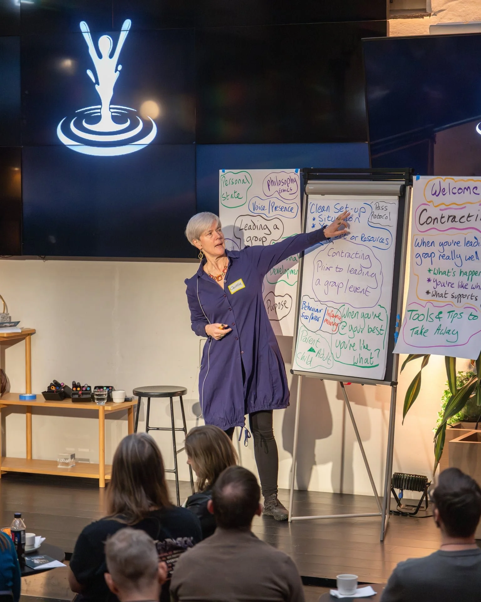 Kursholder Caitlin står på scenen og underviser om Clean Setup. Foran på bildet er bakhodene til deltakerne. Foto. 