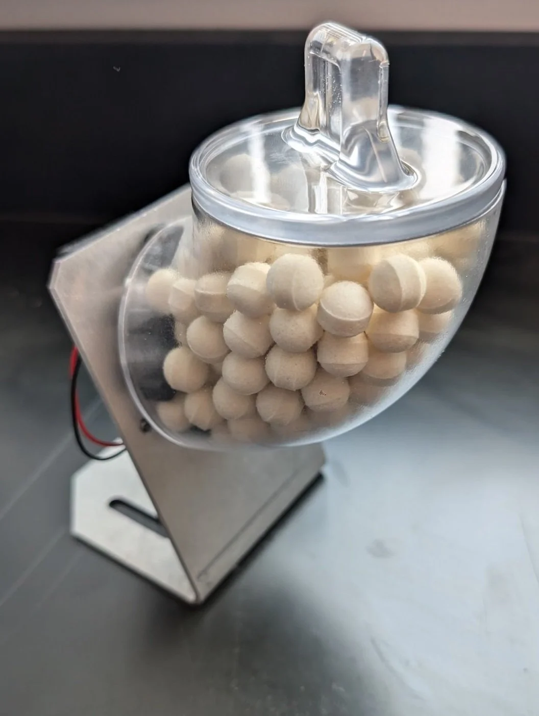 A pellet dispenser, made of a metal bracket and clear hopper filled with white food pellets.
