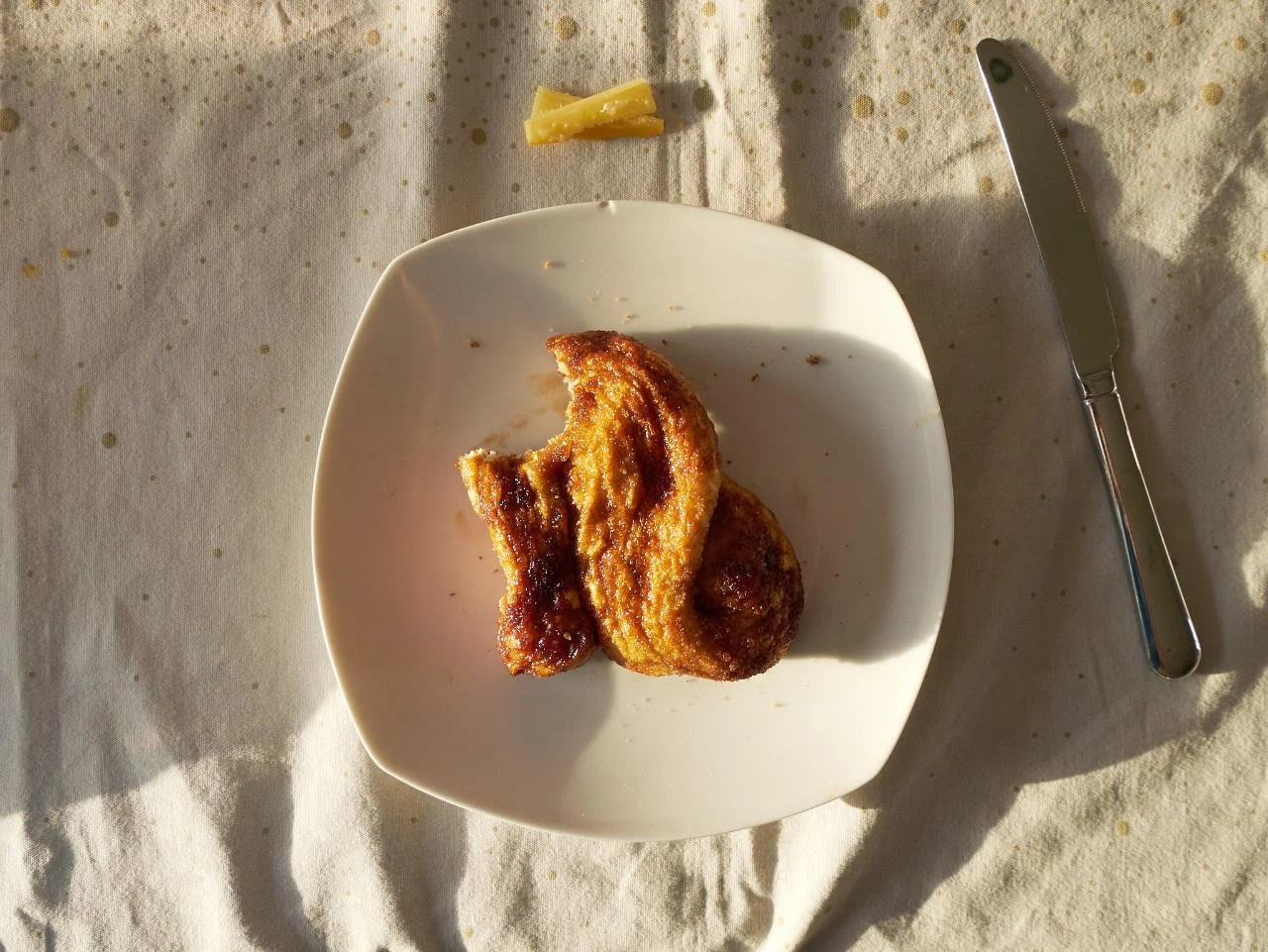 A sugary snack with bit from it sitting on a white plate on a speckled tablecloth. Next to it is a knife and some cheese rinds.