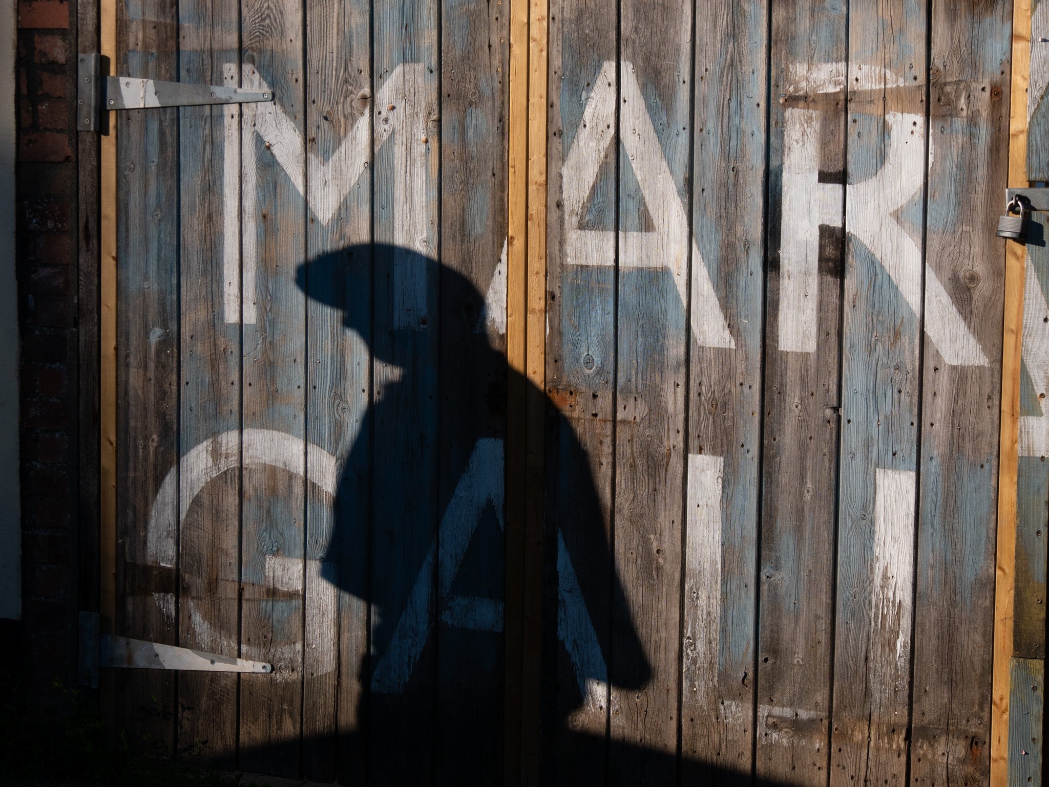 Portrait of a shadow of a man wearing a flat cap against a wooden fence with the letters MAR GALL in large white type.