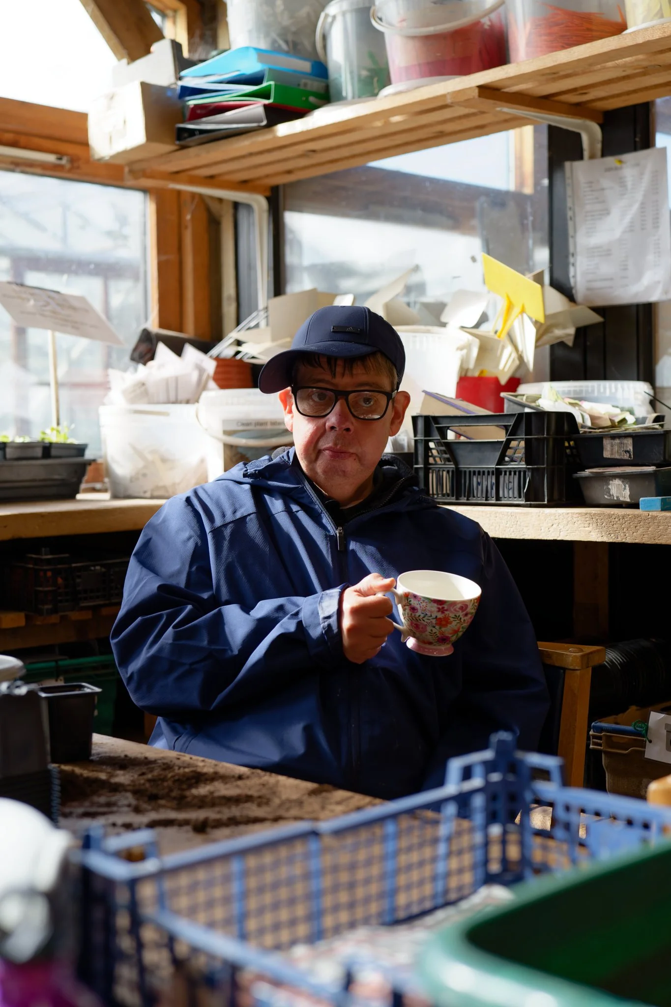 Portrait photograph of a man in a blue hat and jacket sitting in a potting shed and drinking tea from a floral cup.