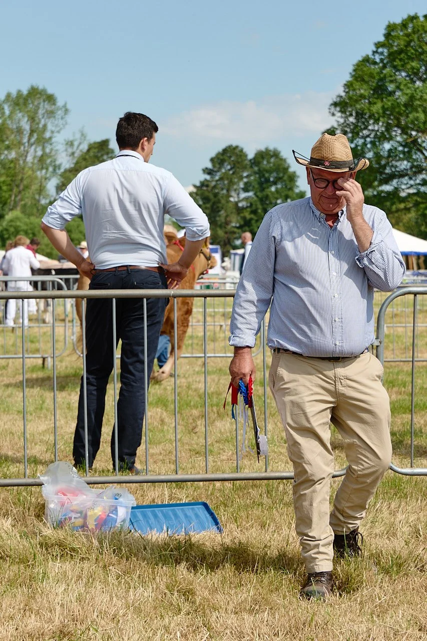 A man in a straw hat adjusts his glasses while he walks away from the judging pens at the North Yorkshire County Agricultural Show. He is holding a clipboard and rosettes.
