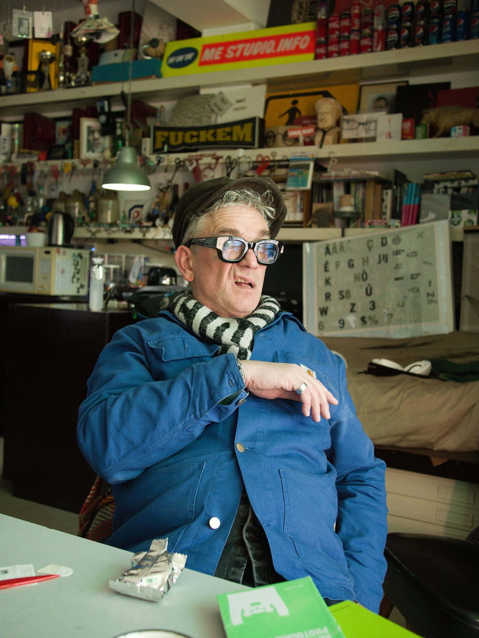 Portrait photo of designer Martin Pyper reacting in mock horror to having his photo taken. He wears a blue chore coat and oversized glasses.