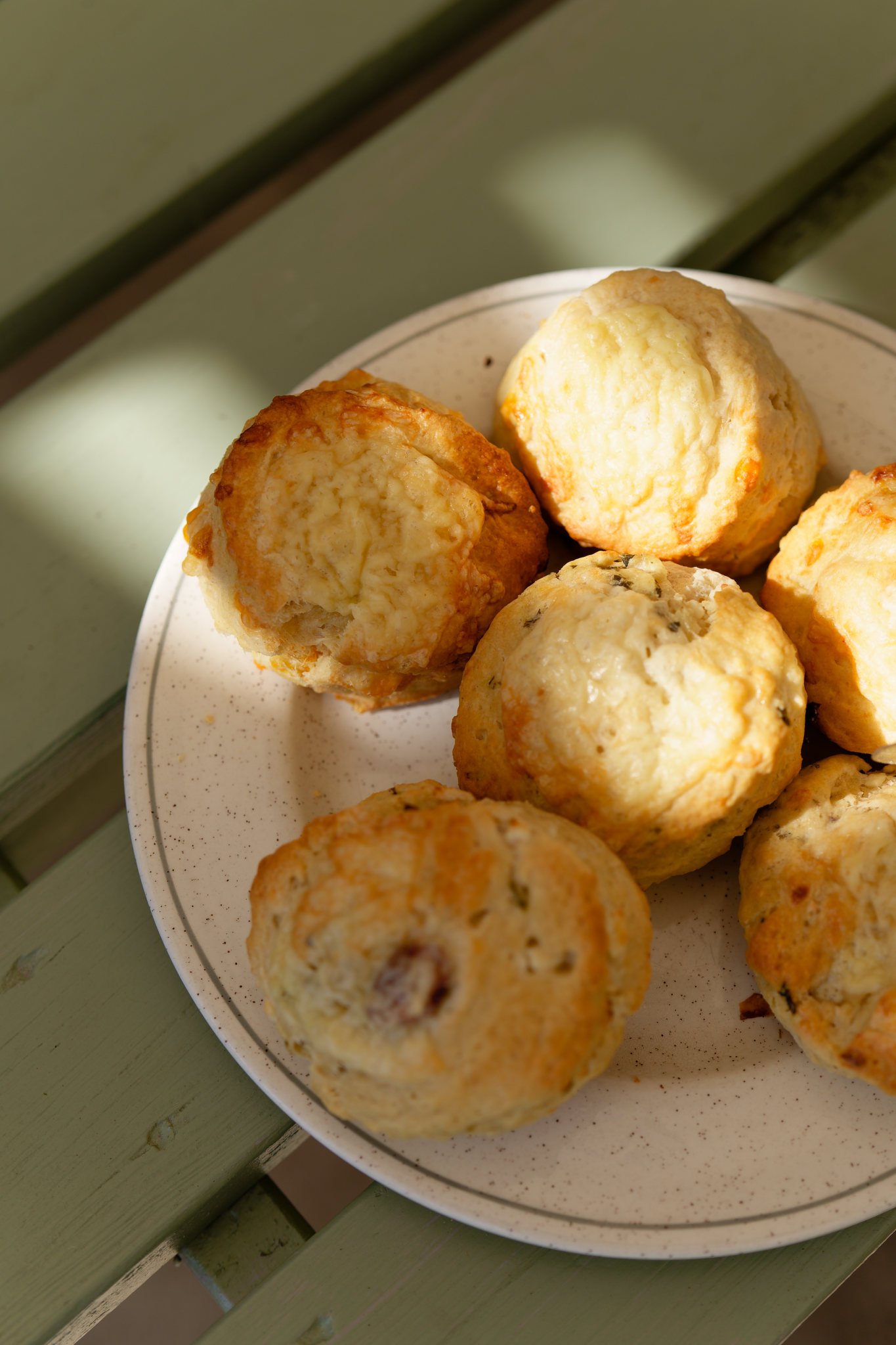 A  plate of six freshly made scones on white place sitting on a light green picnic bench.