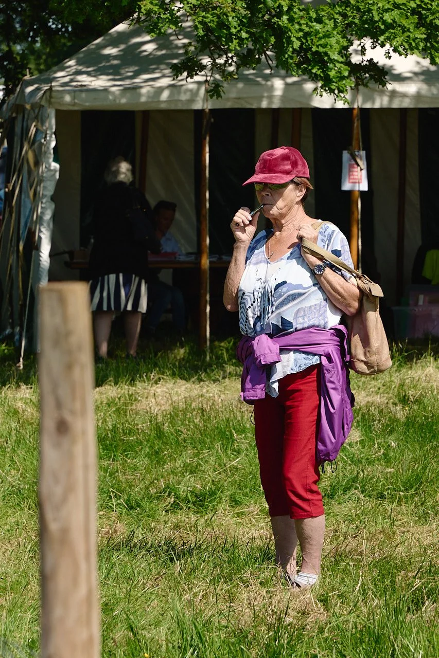 A woman in a red velvet cap and sunglasses with a purple jacket wrapped round her waist vapes while watching the sheep competition.