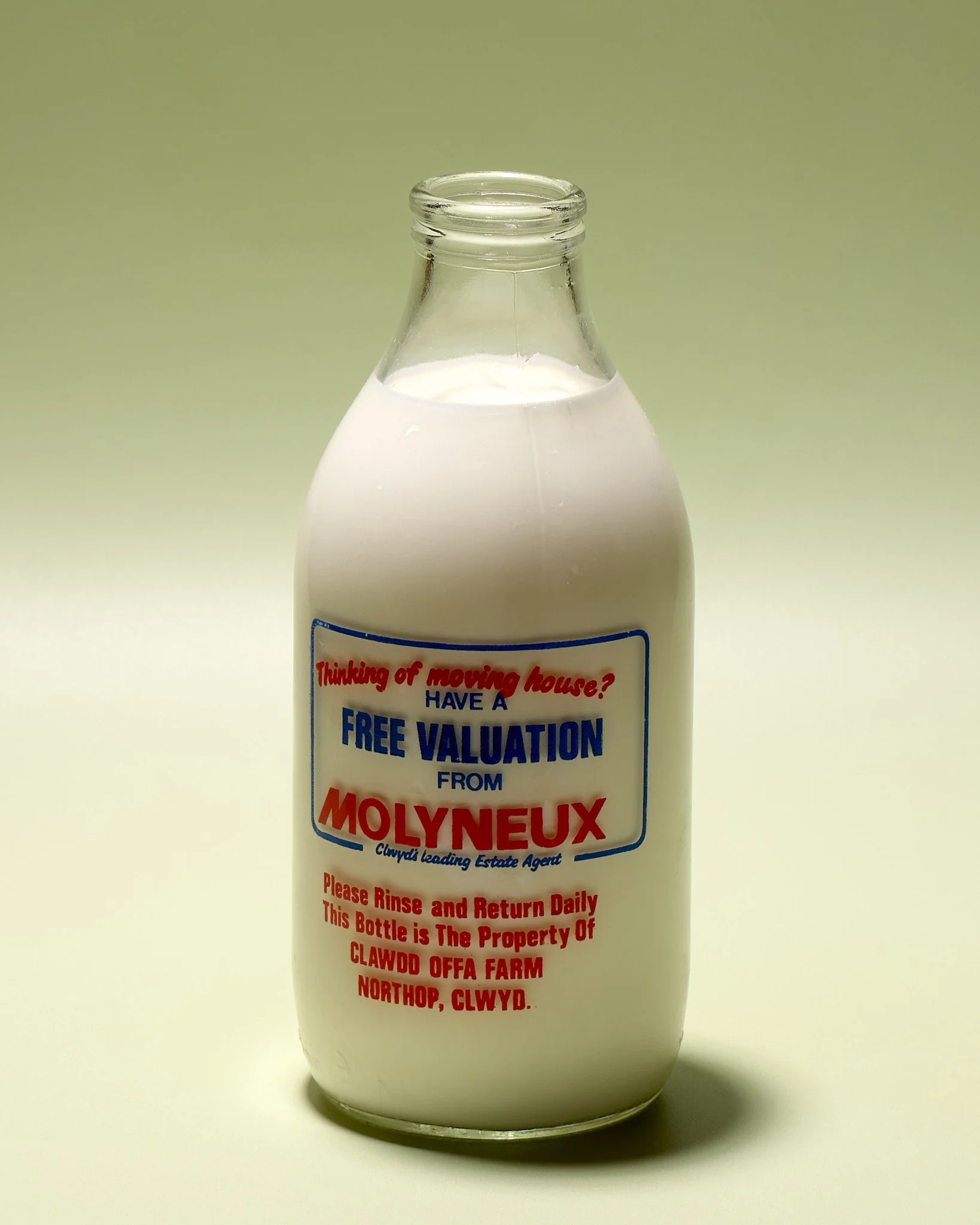 A milk bottle filled with milk shown against a light green gradient background. The bottle has a red and blue printed ad for Molyneux's Estate Agency on it.