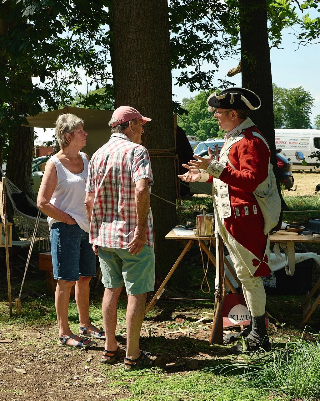 A historical reenactor in a long red jacket with rifle chats to a couple in the shade of the trees.