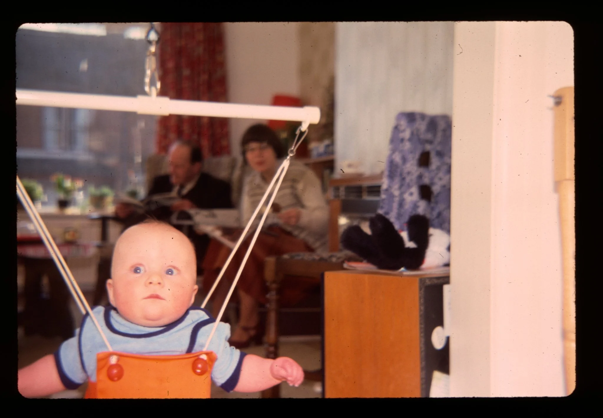 Slide scan of an alarmed looking baby in a bouncer reacting to a camera flash going off. Two people are sat in the background reading.