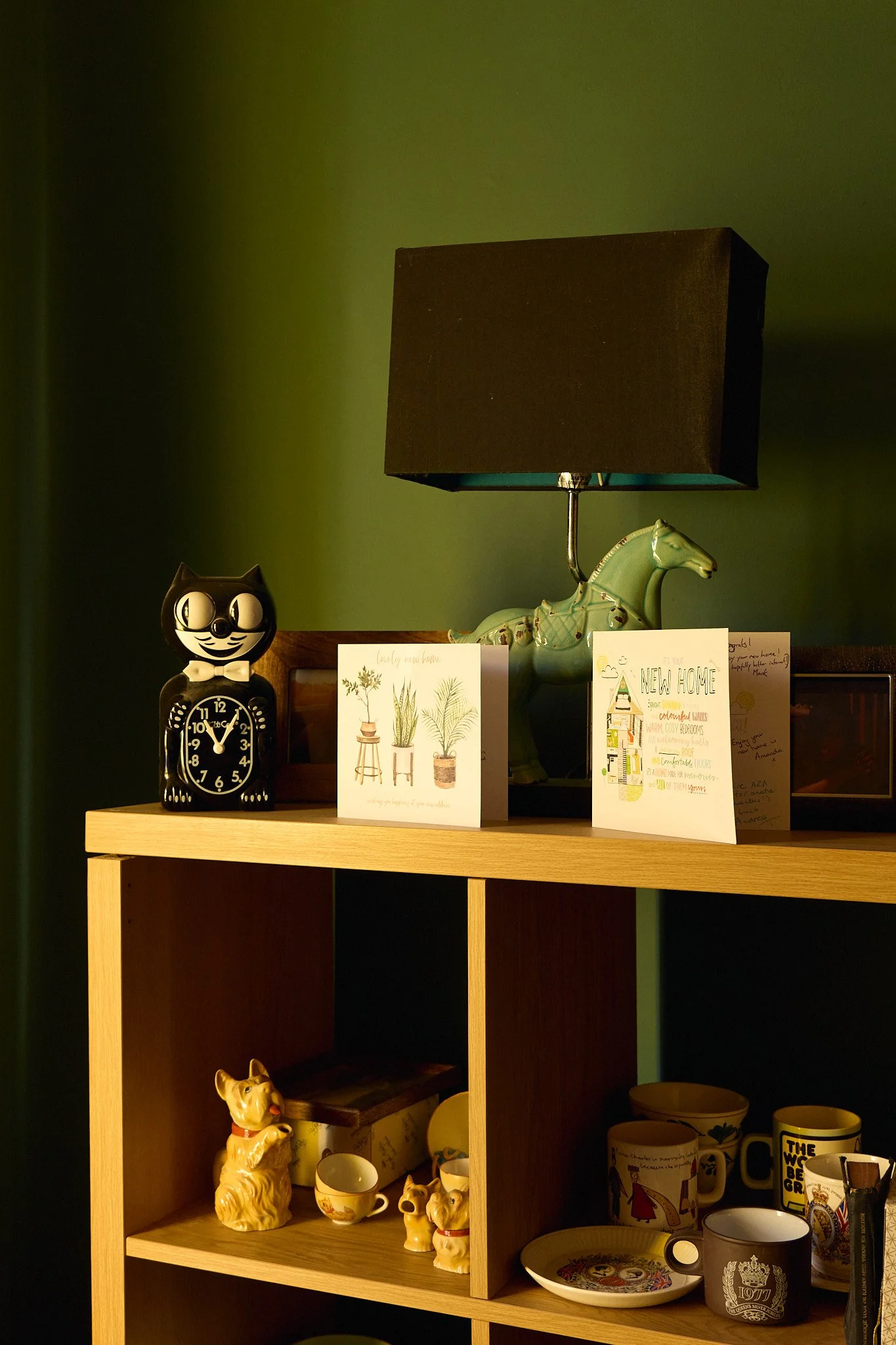 Close up of a bookshelf in a green painted front room with New Home cards. On the shelves are a Kit Kat clock, horse lamp, a begging dog milk jug and various decorative mugs.