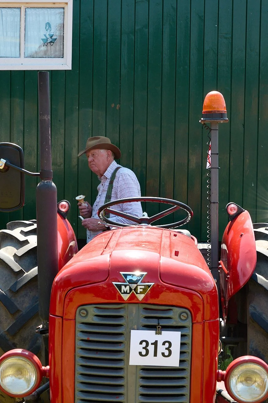 A man in a brown hat eats an ice cream in the shade next to a bright red vintage tractor.