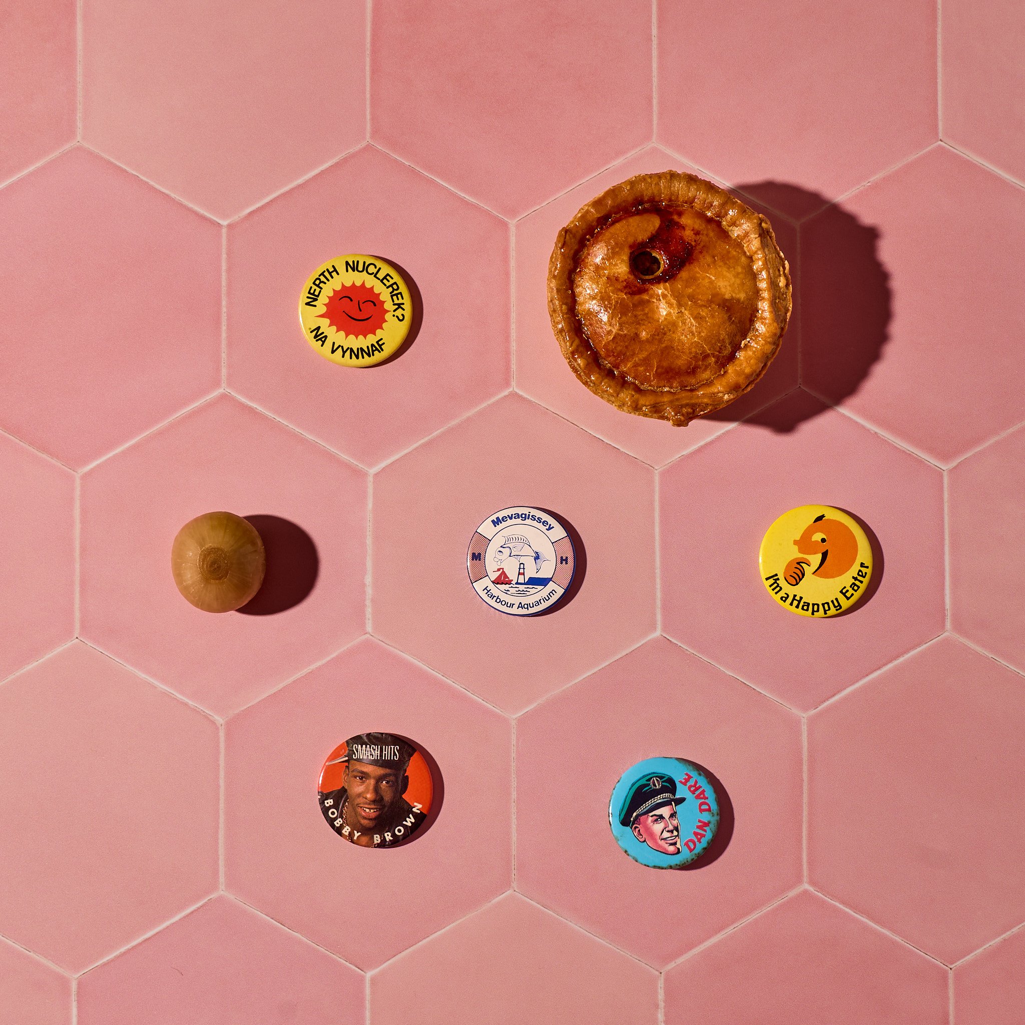 Six colourful badges with various designs arranged on a pink hexagonal tile surface, along with a pork pie and a pickle.