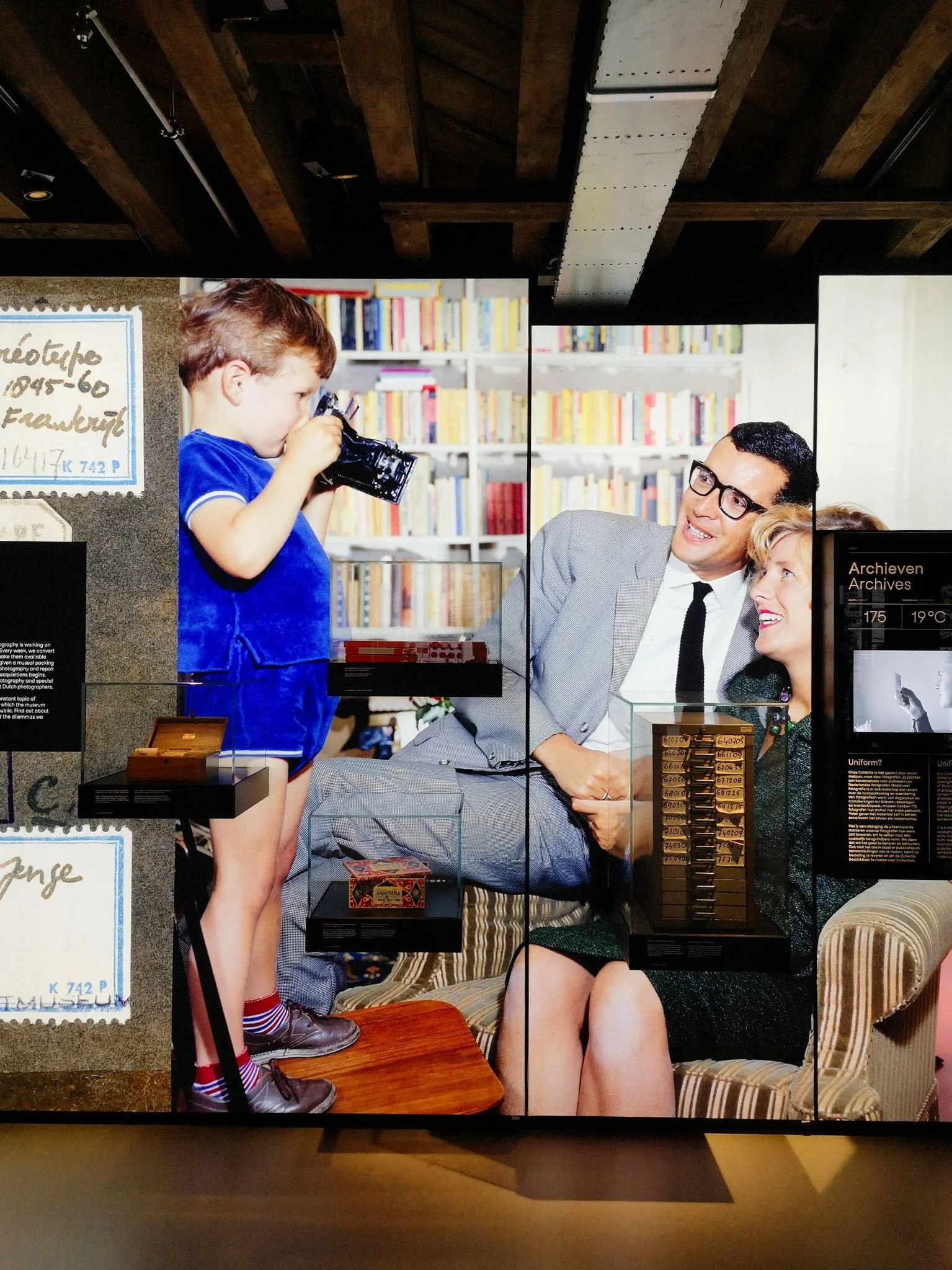 Photo wall featuring displays of photography objects like print and slide boxes. On the wall is a panelled image of a child taking a picture of (presumably) his parents.
