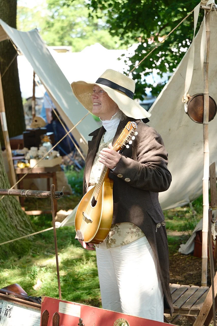 A reenactor in long brown jacket and straw hat holding a round string instrument while chatting to people.