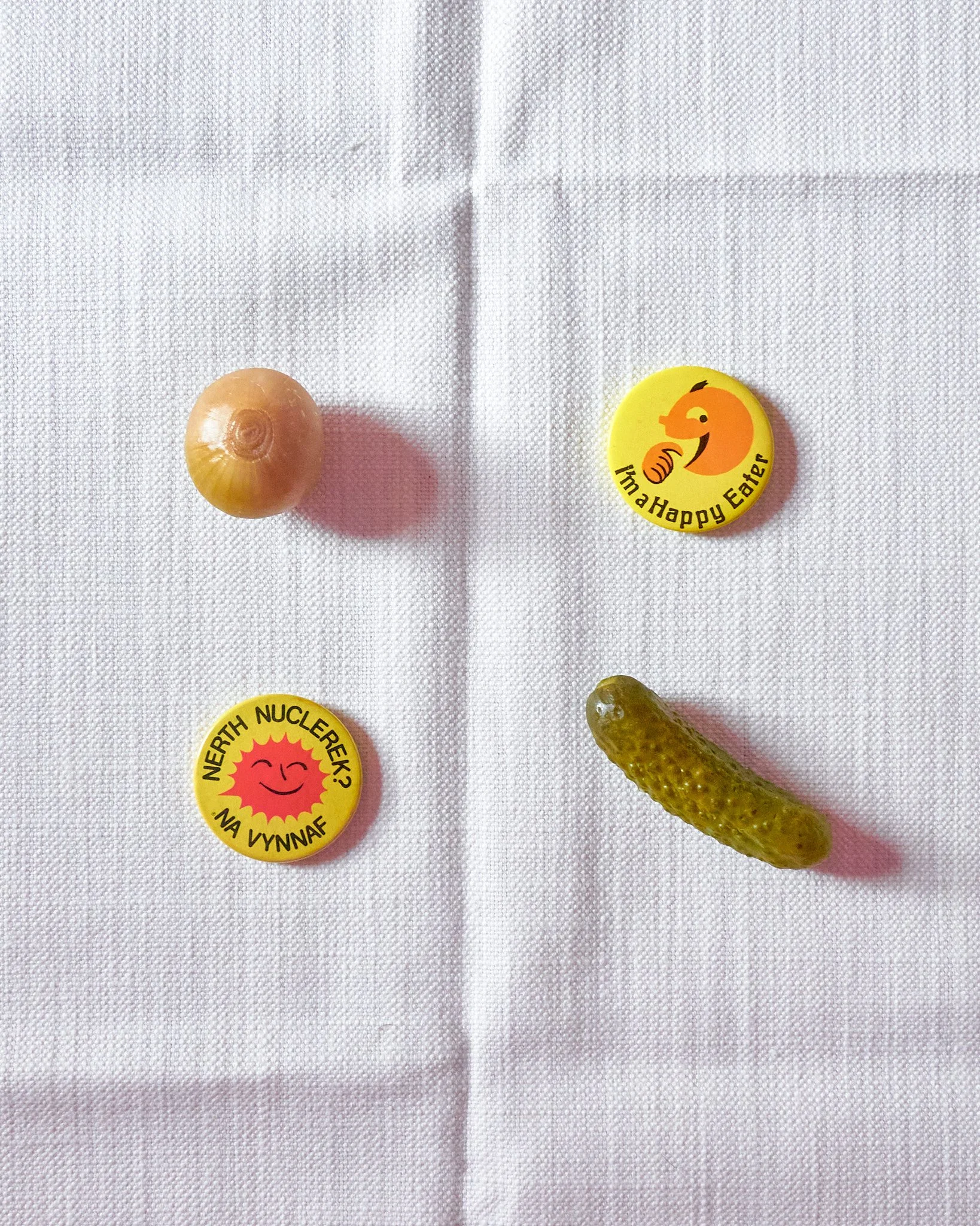 A white cloth with two pins featuring cartoon faces and messages, a small onion, and a small pickle.