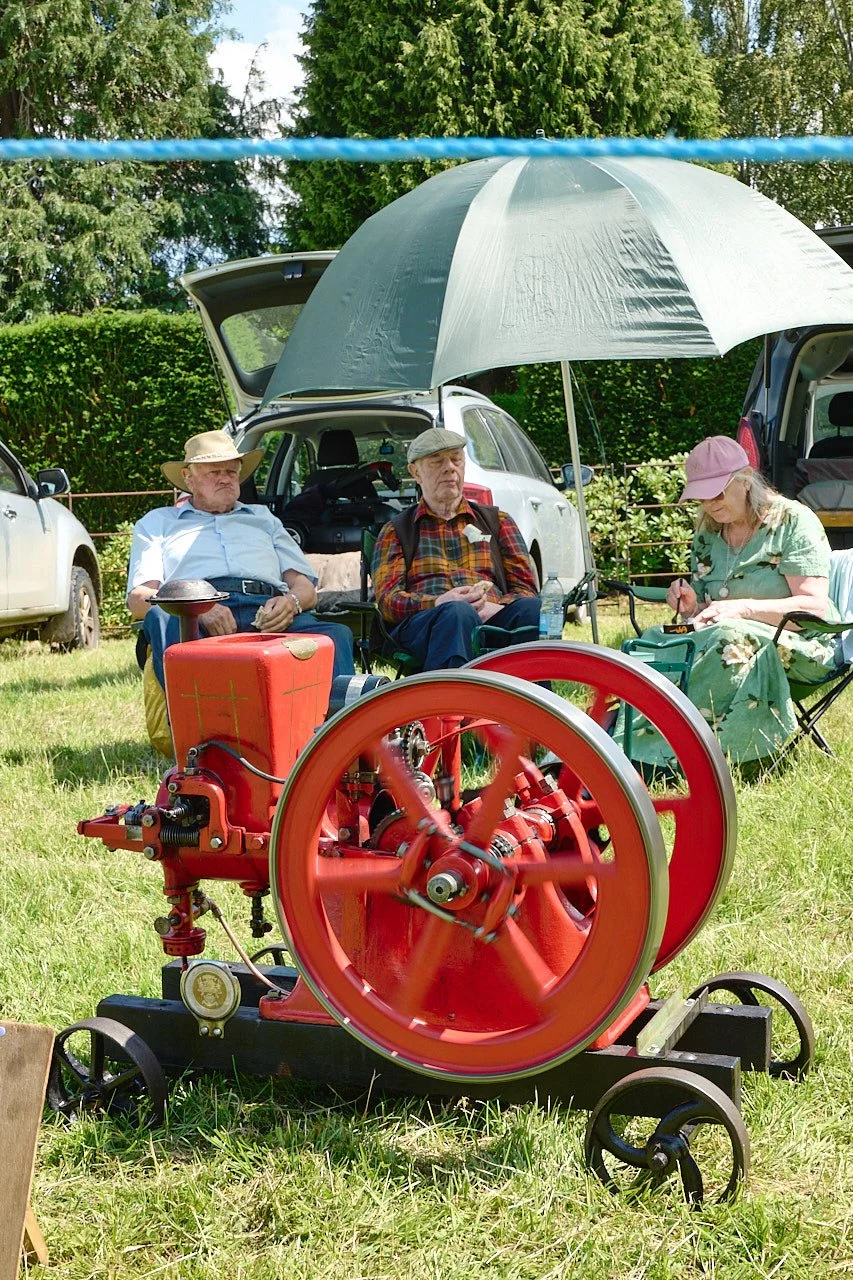 Three people sitting in the shade of a big green umbrella eating. They are sitting behind a small red steam engine.