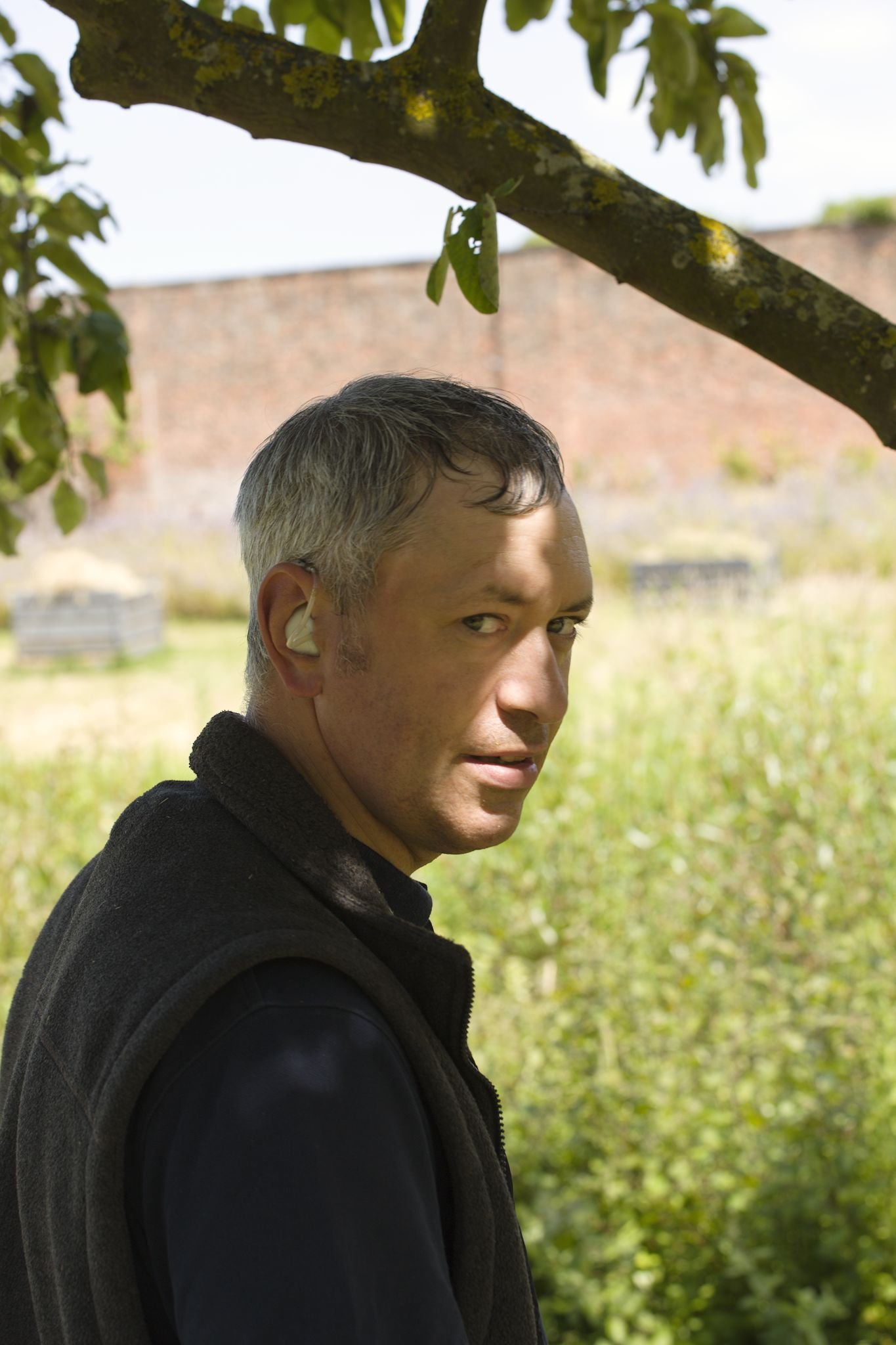 Portrait photo of a man with grey hair standing in the shade of a tree during a hot day. He has a hearing aid and is wearing a black fleece gilet. He is looking over his shoulder at the camera.