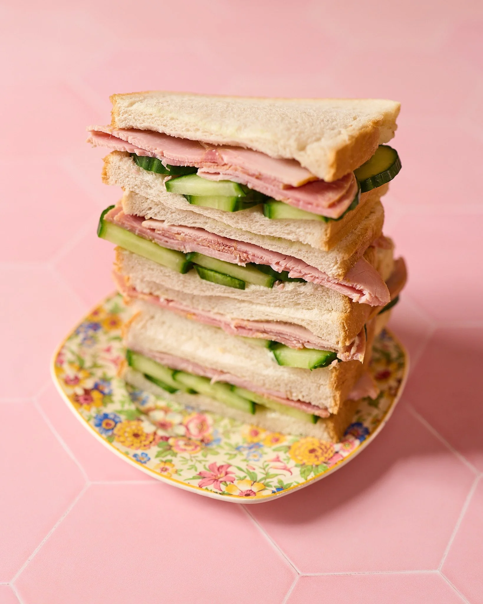 A pile of ham and cucumber sandwiches balanced on top of a flowery patterned yellow plate. The background is pink hexagonal tiles.