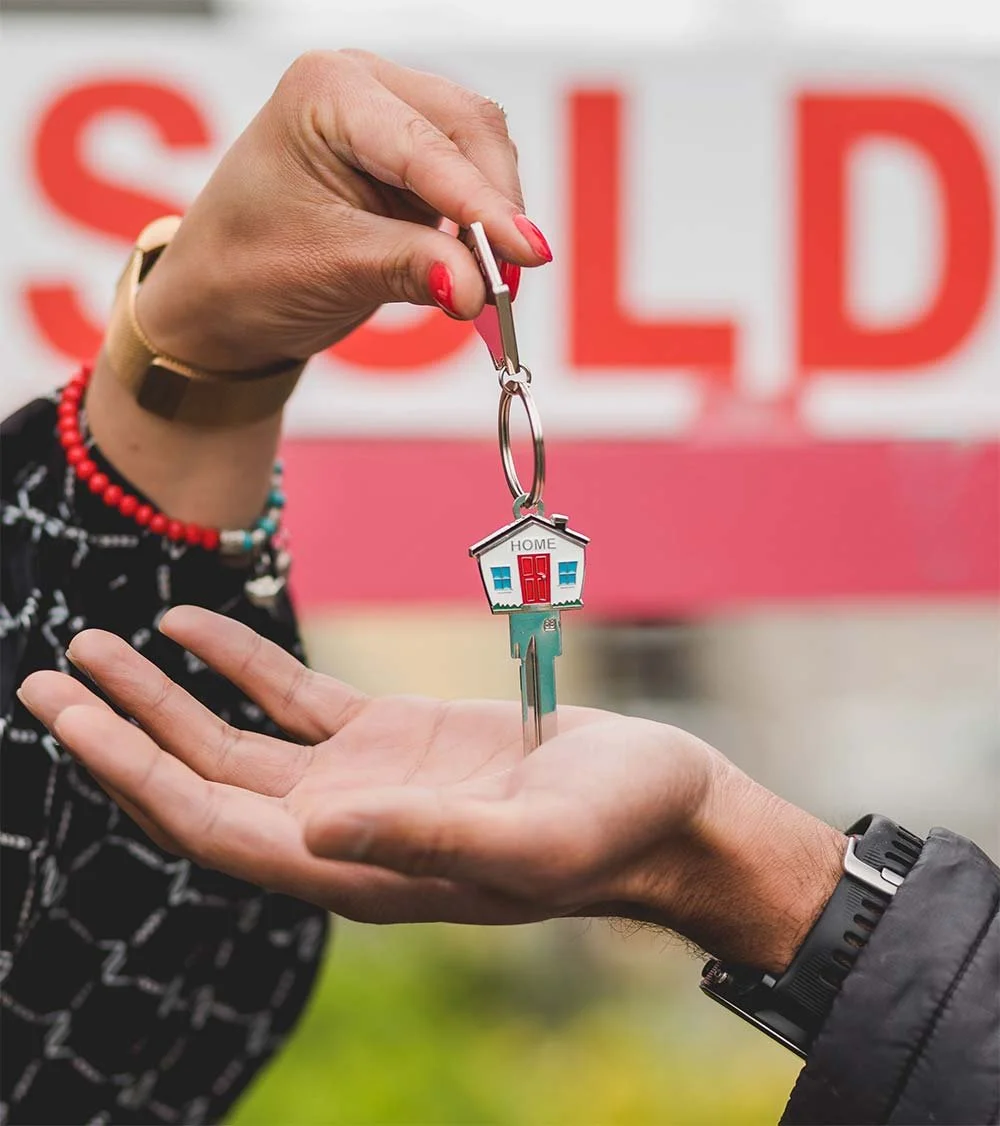 Person handing a key to another person in front of a "Sold" house sign.