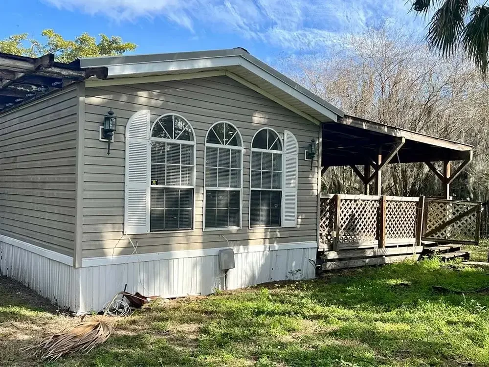 Single-story beige home with white trim, three arched front windows, and a covered wooden porch, surrounded by grass and trees.