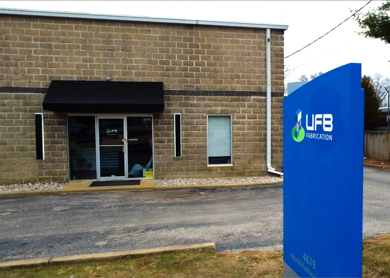 Exterior view of a brick building with a black awning over the entrance and a blue sign that reads 'UFB Fabrication' outside.