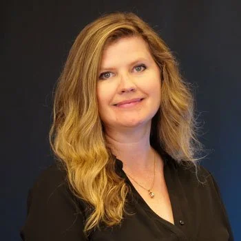A woman with long wavy blonde hair wearing a black top, smiling against a dark background.