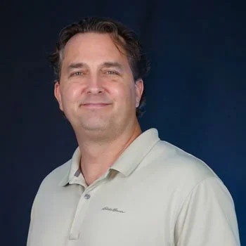 Portrait of a man with short dark hair wearing a light-colored polo shirt, smiling, against a dark background.
