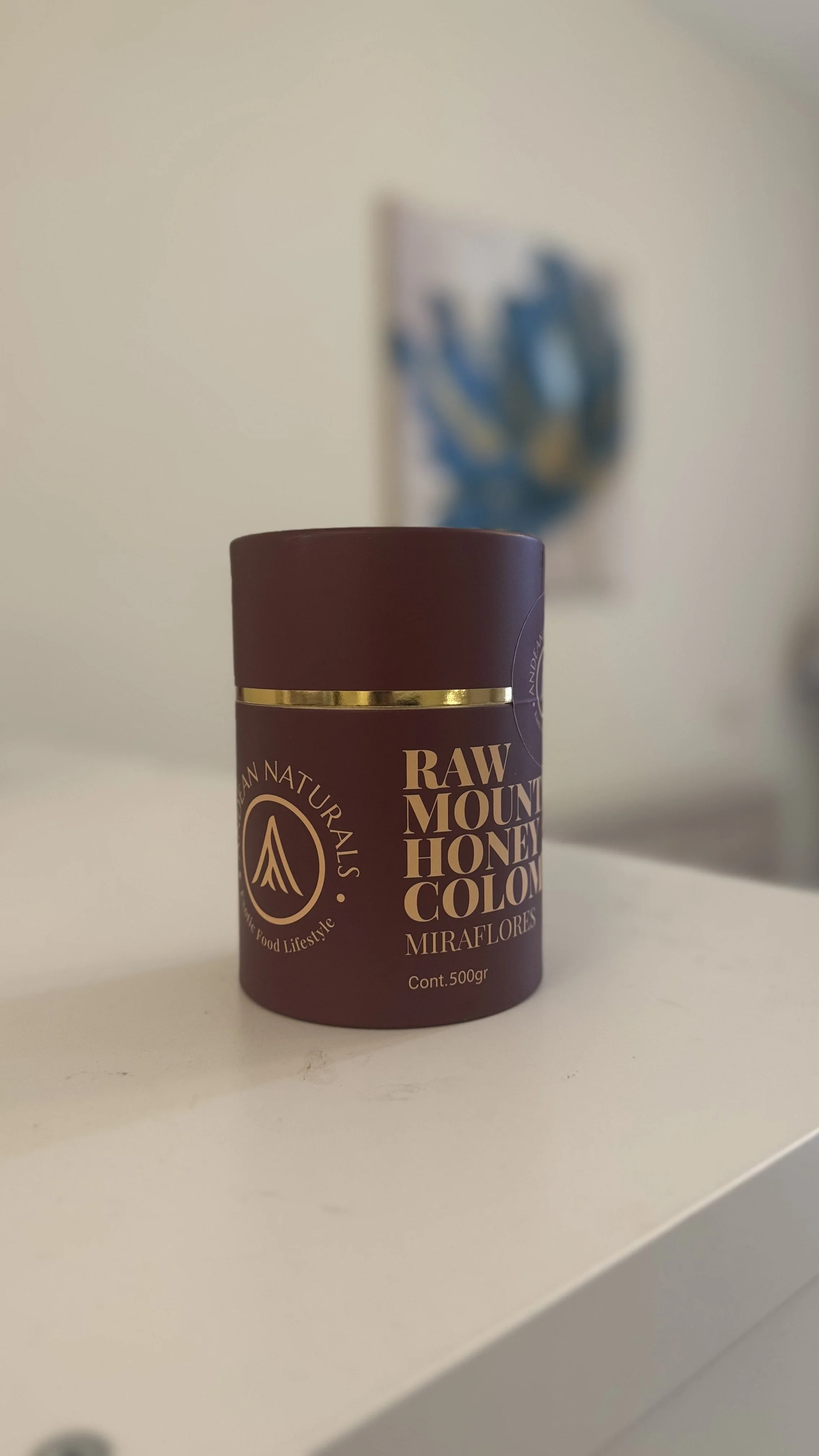 A cylindrical container of raw mountain honey colon from Miraflores placed on a white surface with a blurry wall and artwork in the background.
