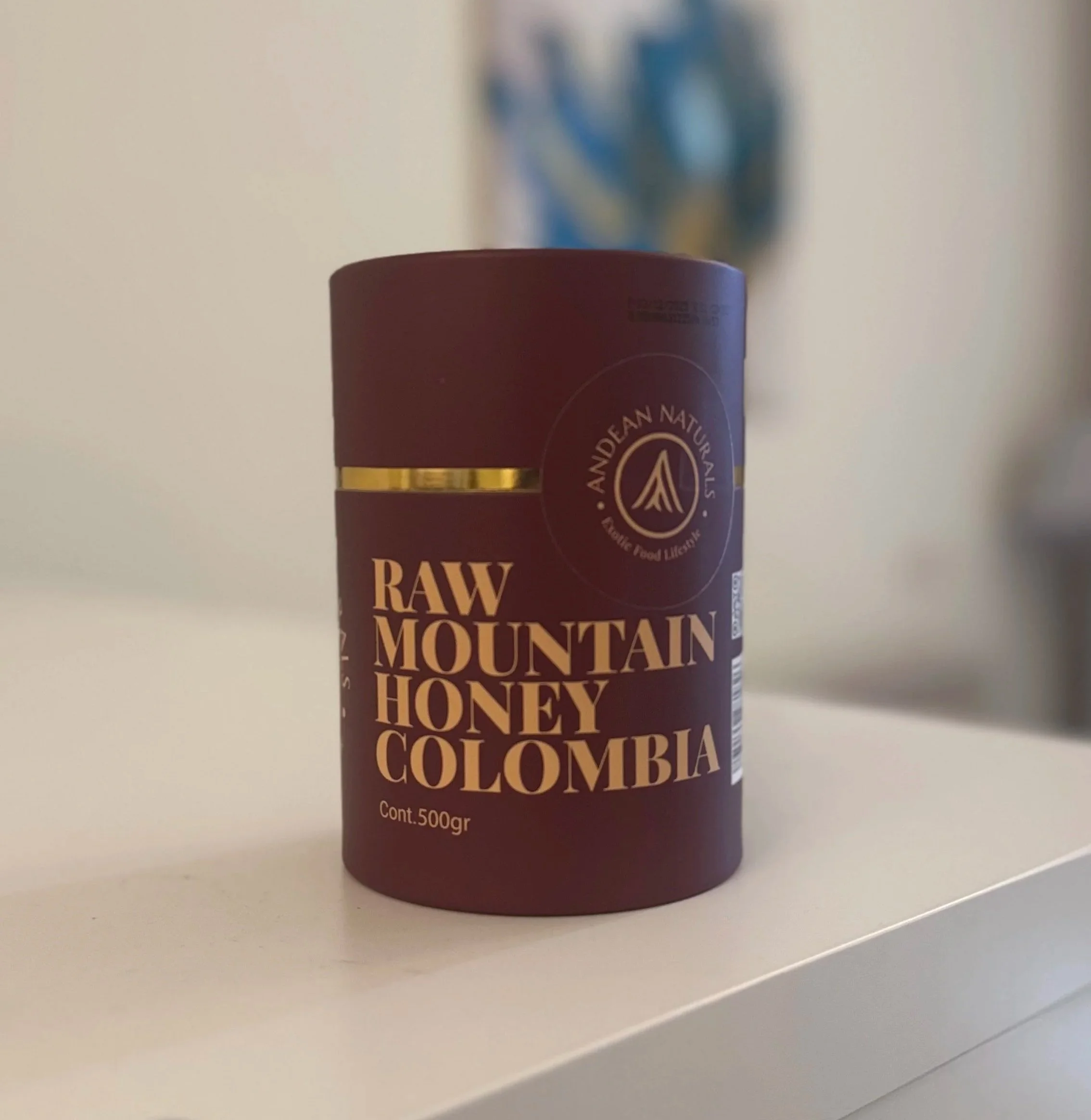 A cylindrical container of raw mountain honey from Colombia with a maroon label and gold accents, placed on a white surface.