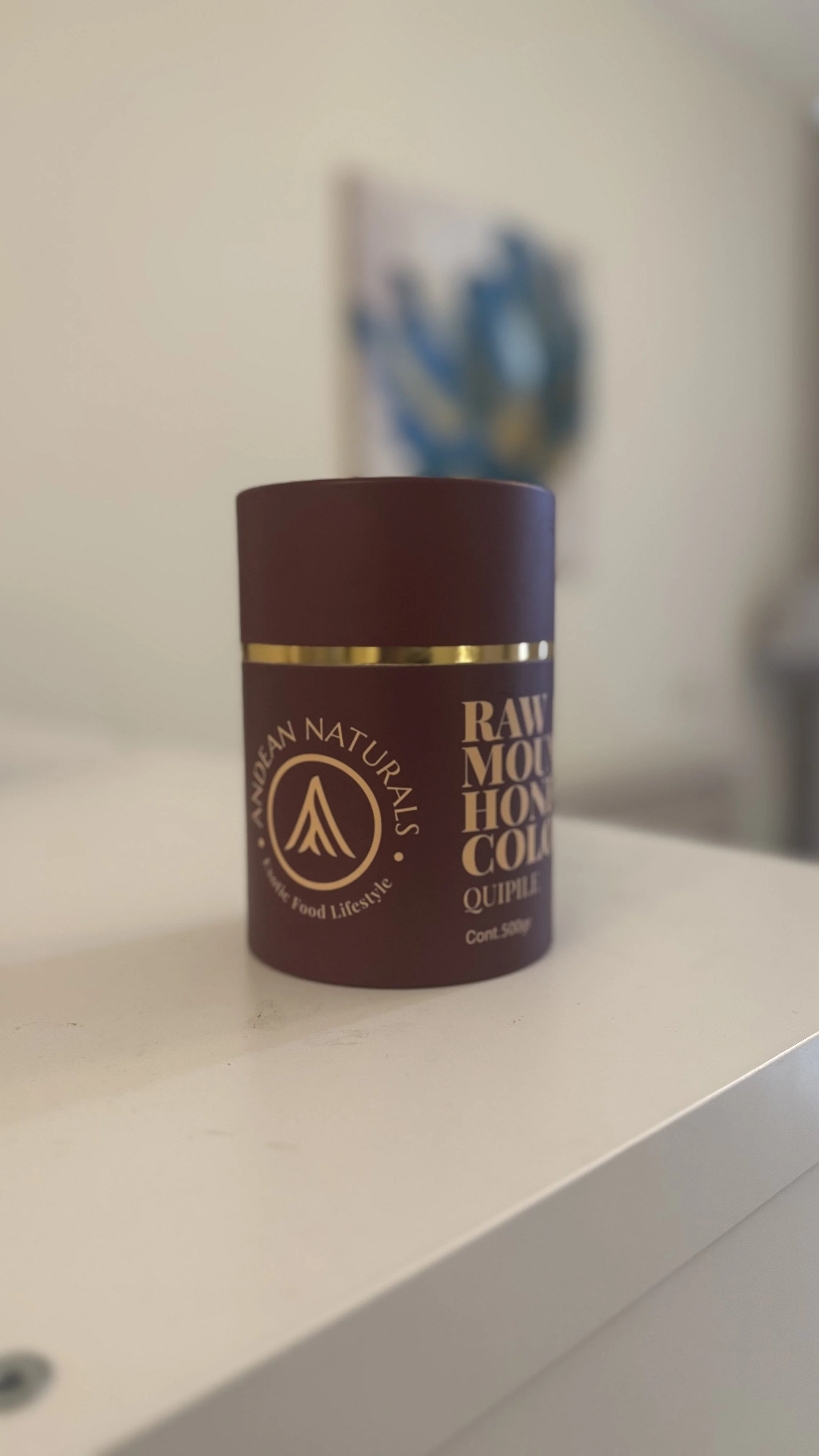 A maroon container labeled 'Raw Mountain Honey Comb' from A Mediterranean Naturals sitting on a white surface with a blurred abstract art piece on the wall in the background.