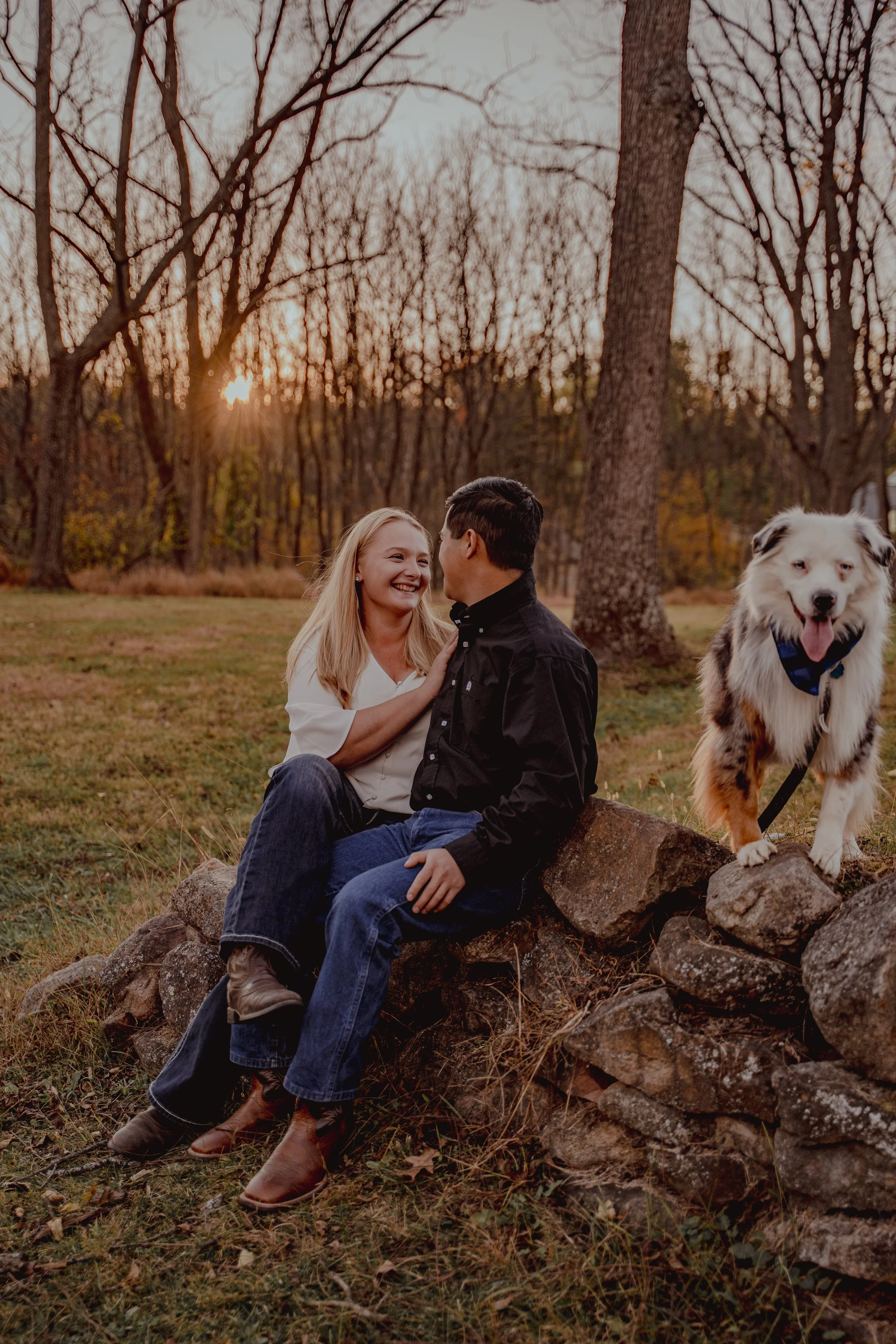 A couple sitting on a stone wall, smiling and looking at each other, with a happy dog standing next to them in a park during sunset.