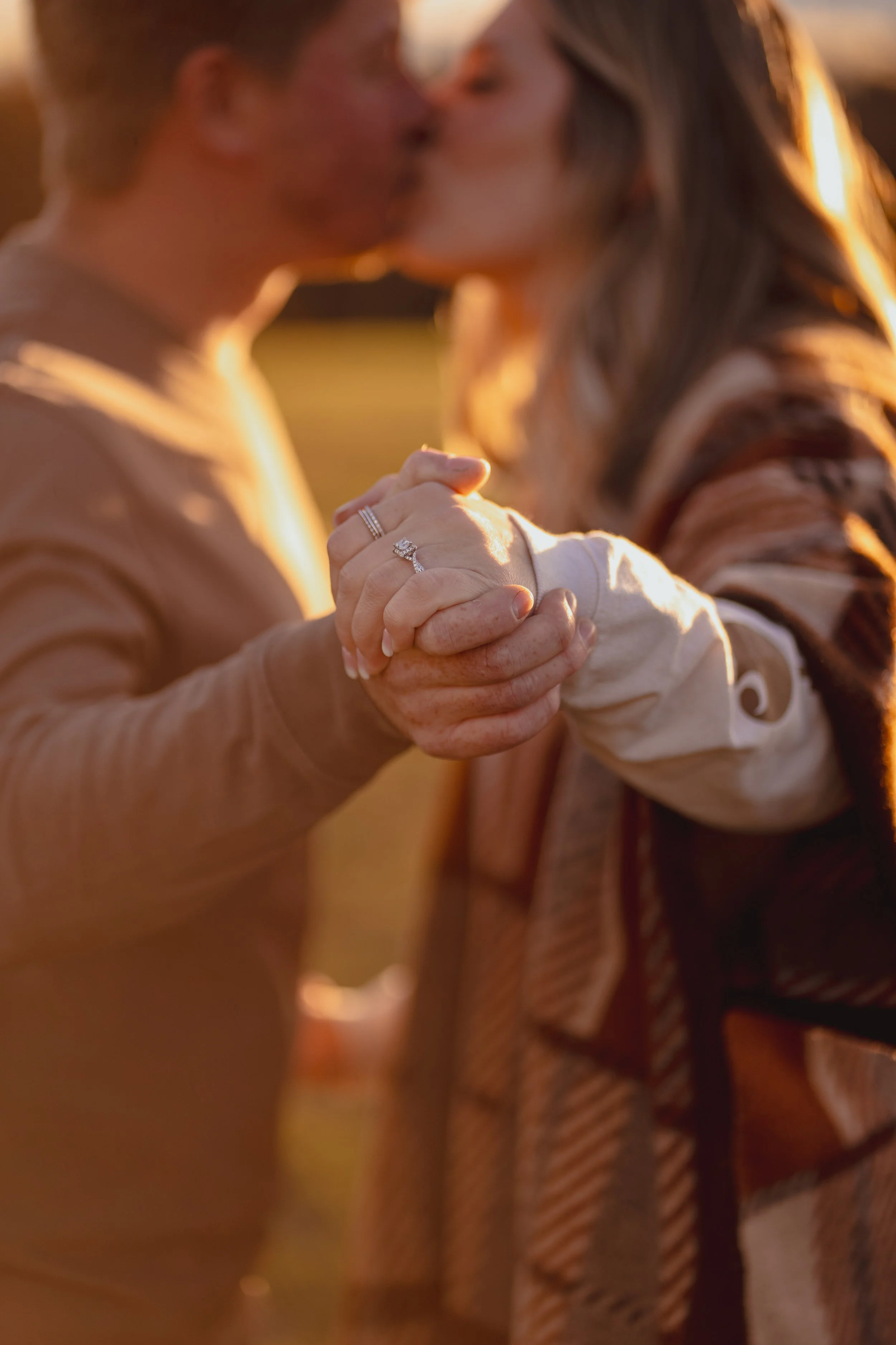 A couple kissing while holding hands, showcasing a wedding ring in warm sunset lighting.