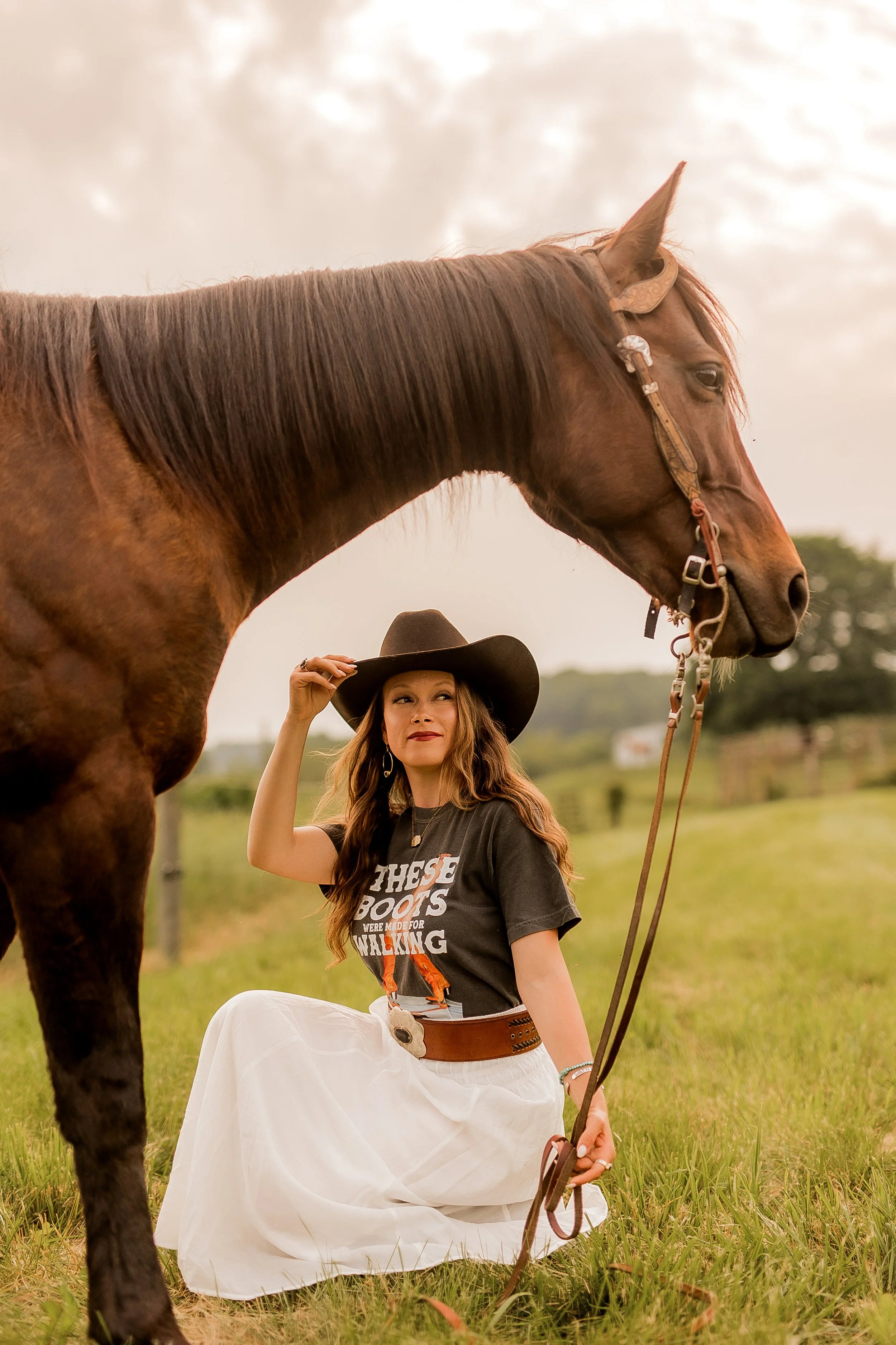 A woman in a black cowboy hat and white skirt kneels on the grass holding a brown horse with a bridle, outdoors on a cloudy day.