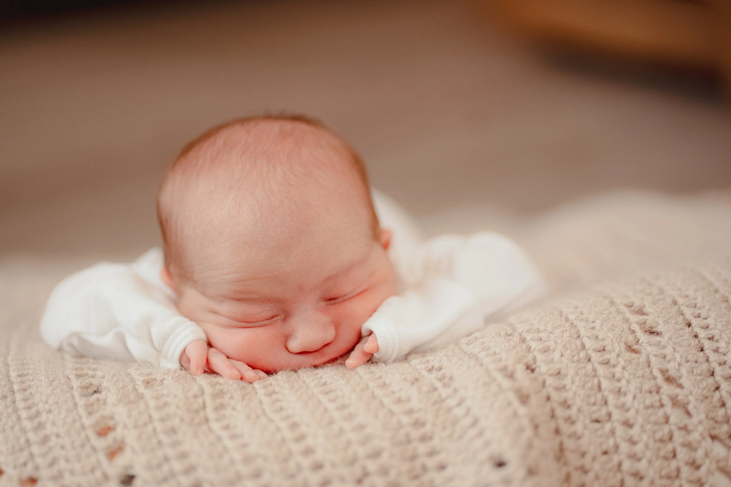 Close-up of a sleeping newborn baby lying on a textured beige blanket, resting with hands near face.