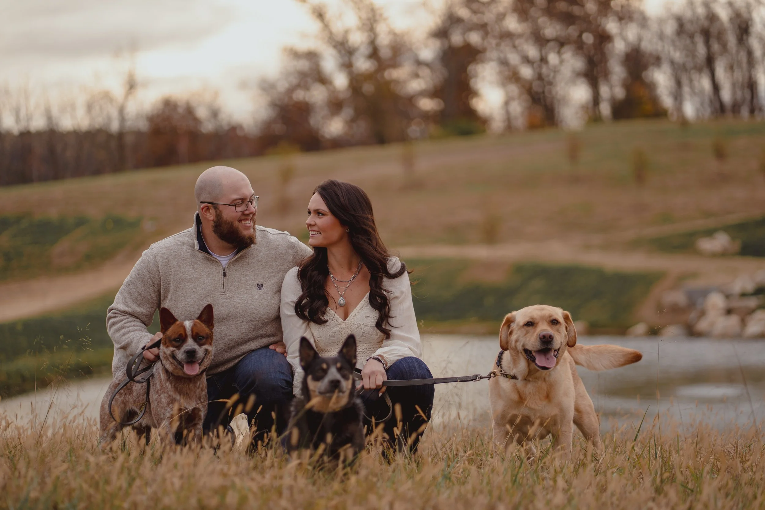 A smiling couple sitting on grass with their three dogs near a lake during autumn, with trees in the background.