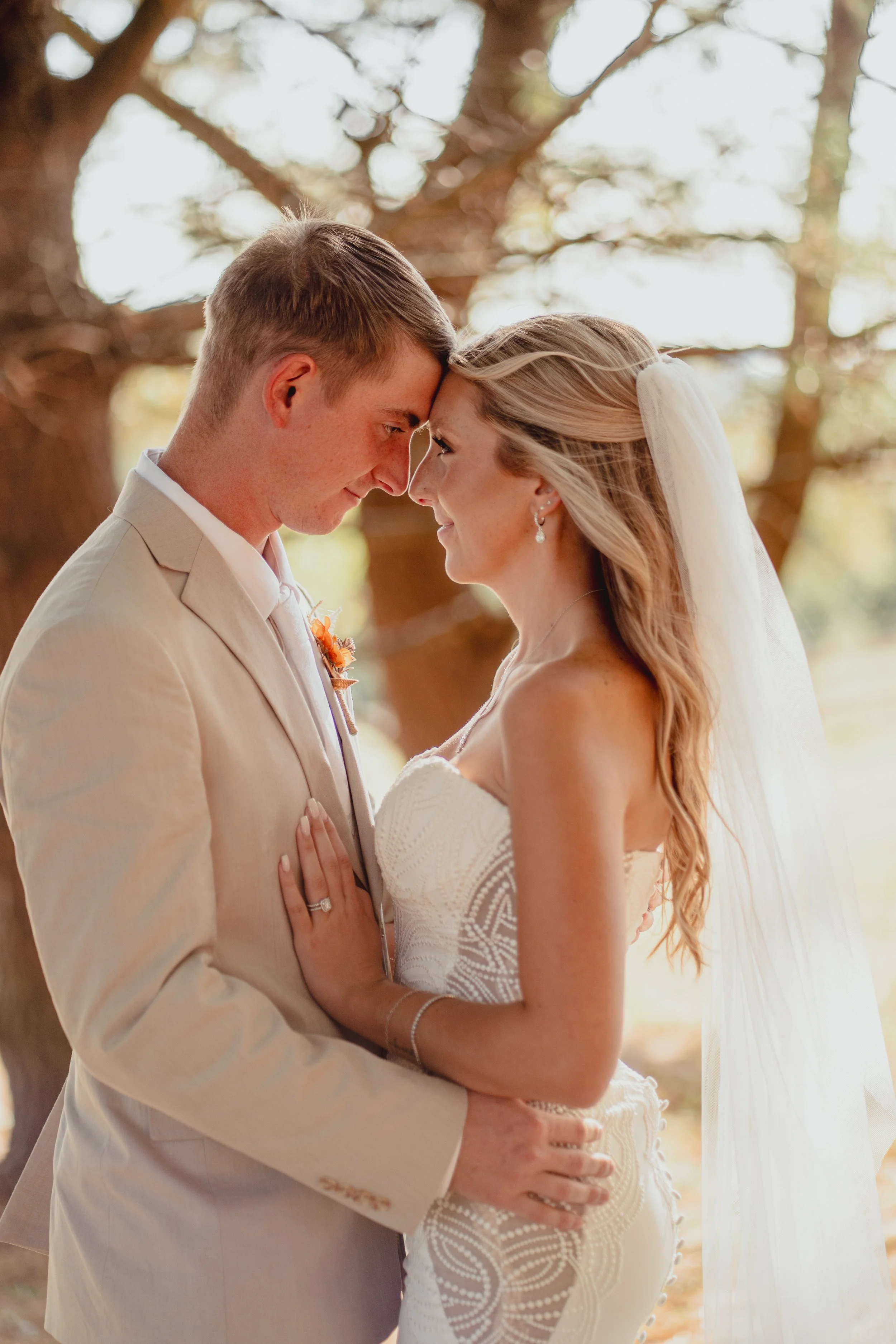 A bride and groom are standing very close with foreheads touching, outdoors under a large tree, during their wedding, with blurred foliage in the background.