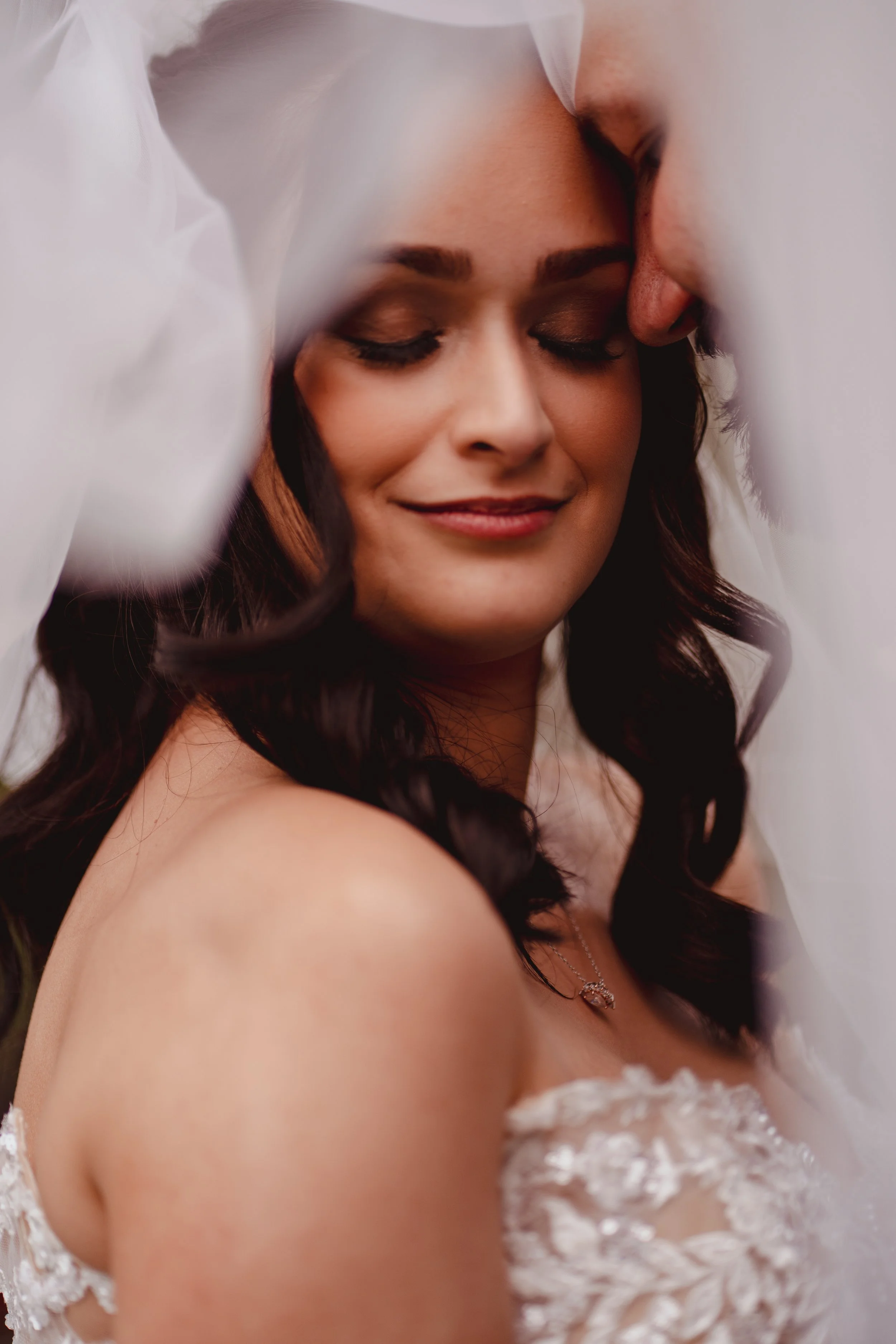 A bride with dark, wavy hair and a wedding dress, smiling with eyes closed, framed by white fabric or veil.