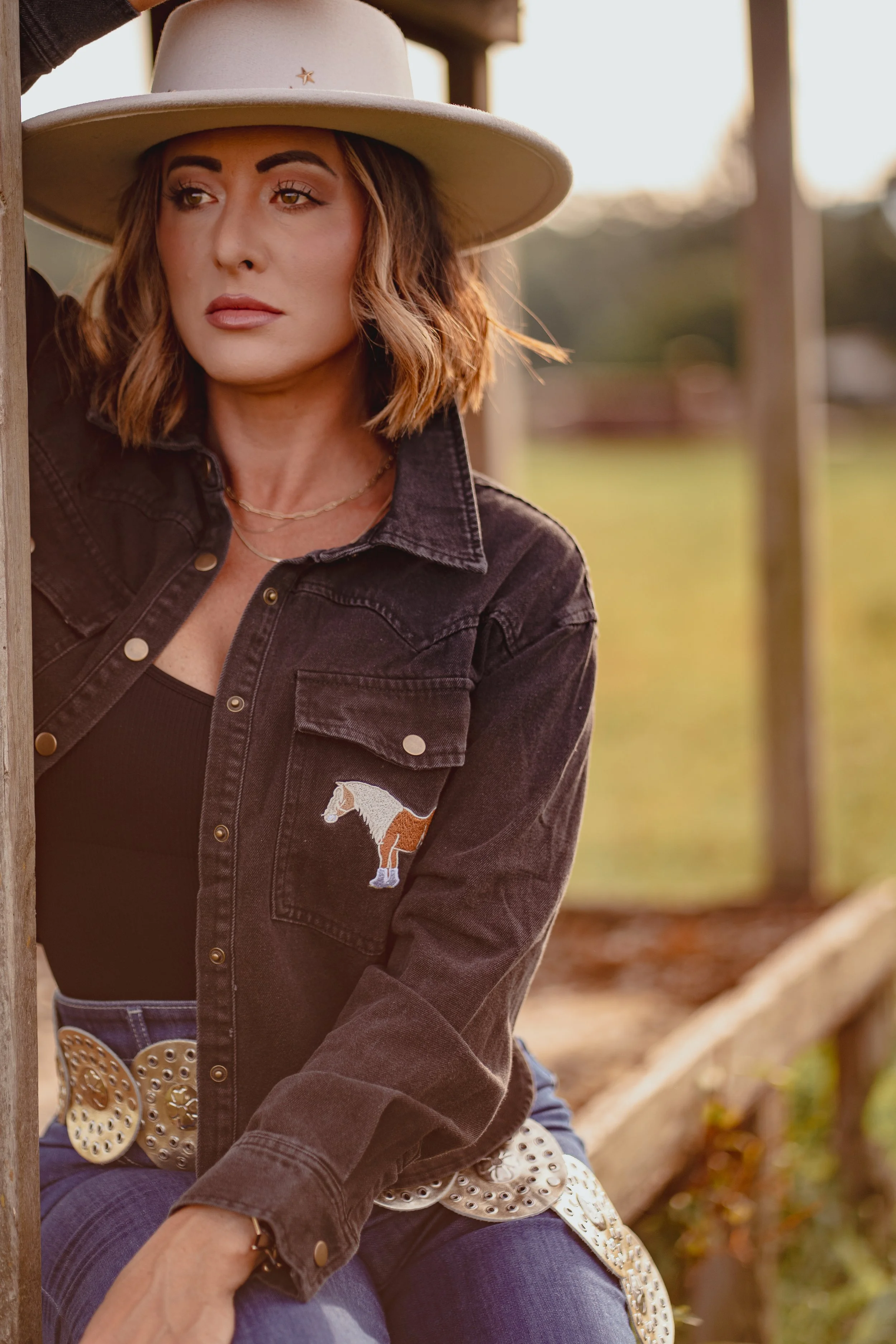 A woman with shoulder-length wavy hair wearing a beige hat with a small star, a black denim jacket with a horse embroidery on the chest, a black top, and decorated belt sitting outdoors near a wooden fence during sunset.