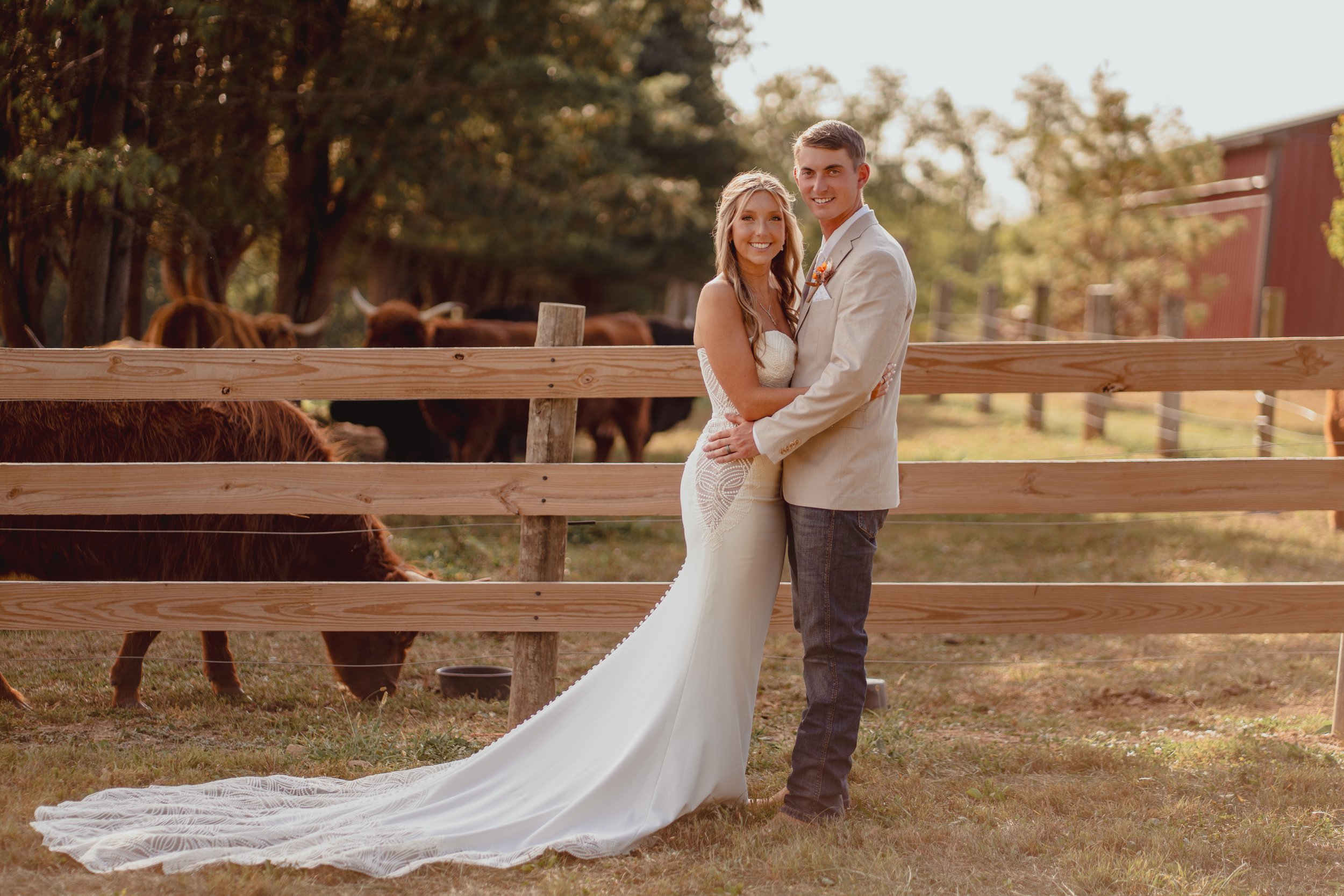 A happy couple in wedding attire standing close together outdoors, with cows and trees in the background, on a farm or rural setting.