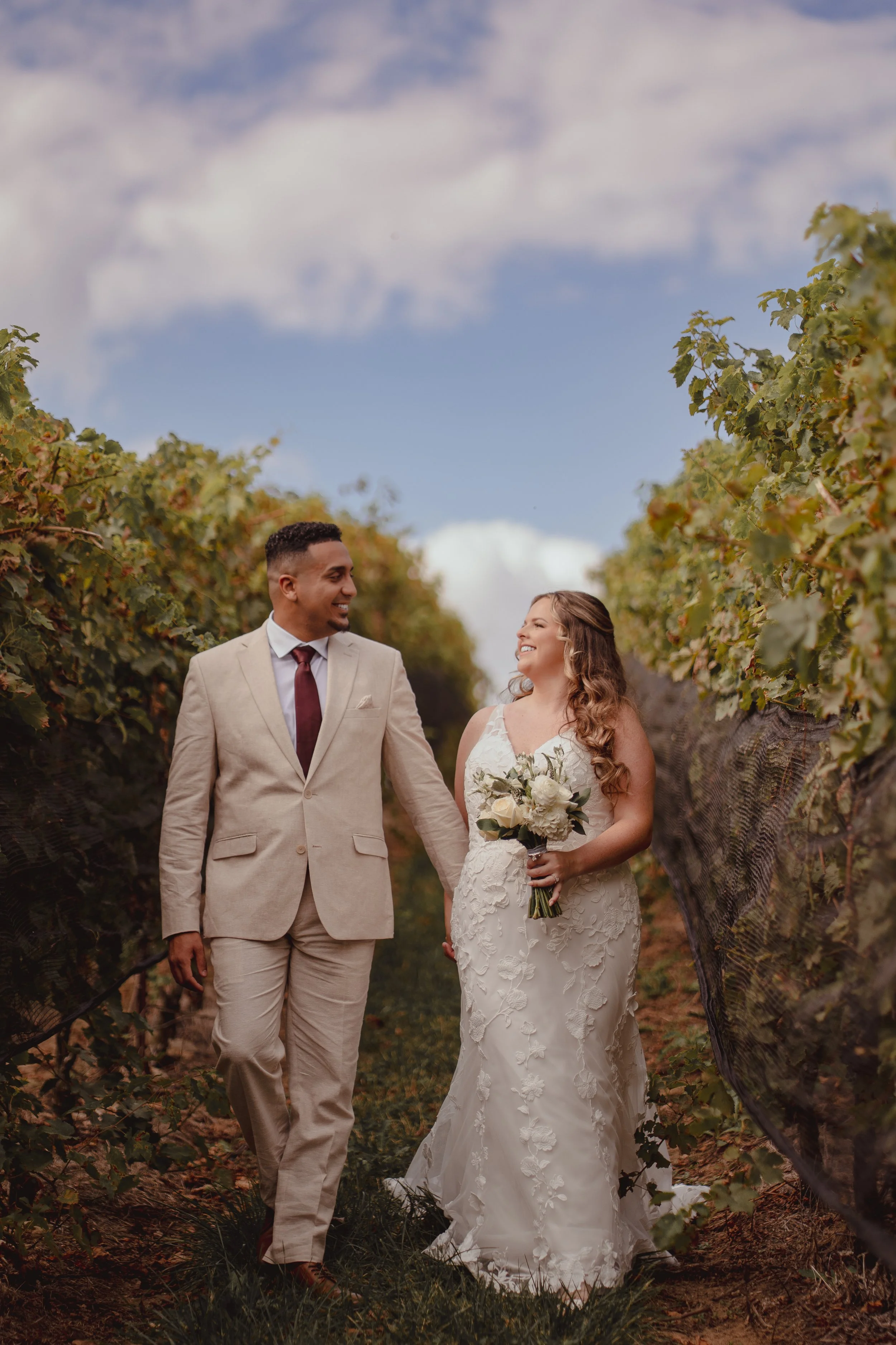 A newlywed couple walking hand-in-hand through a vineyard, smiling at each other, during daytime with a partly cloudy sky.