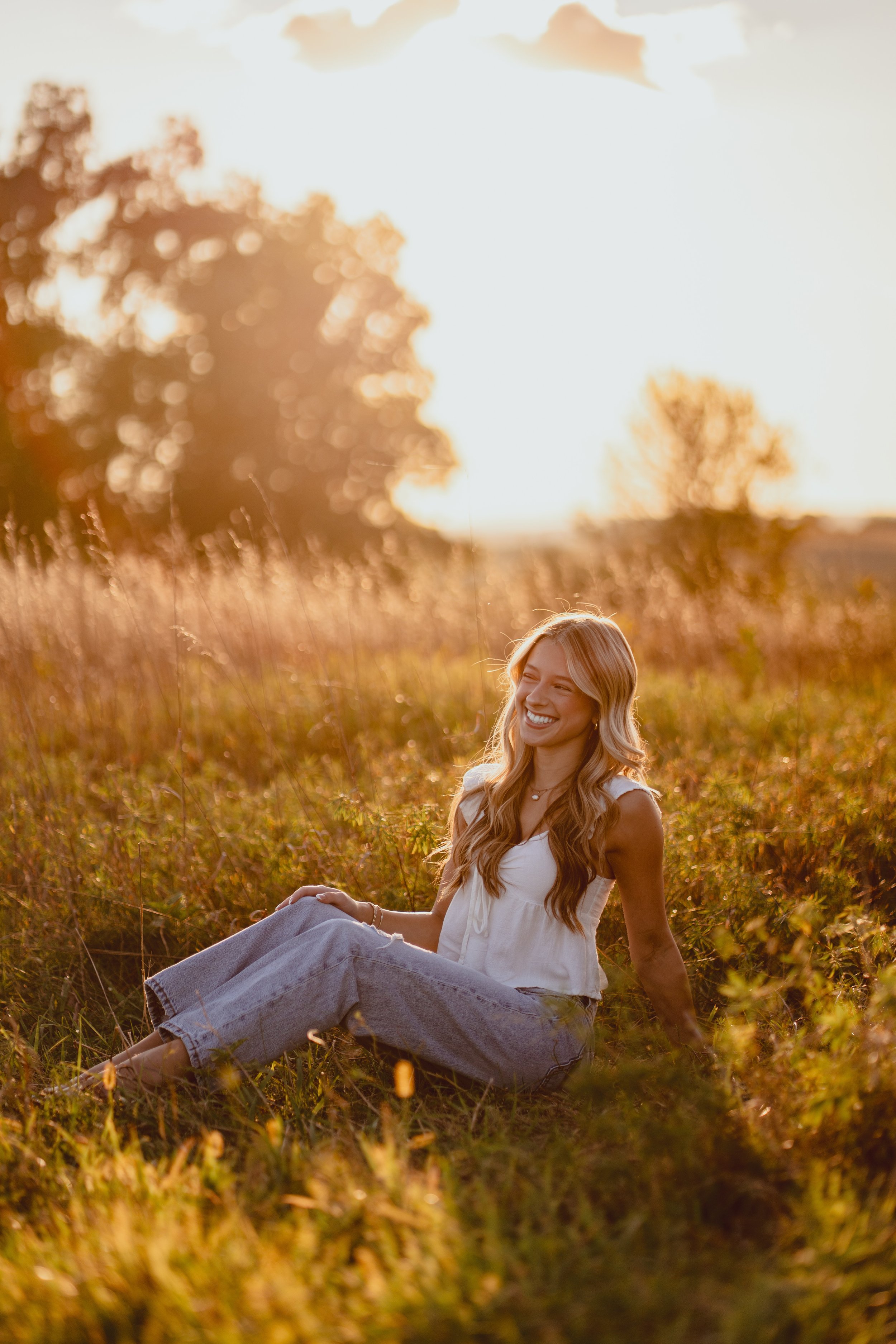 A young woman with long blonde hair smiling and sitting in a field of tall grass during sunset.