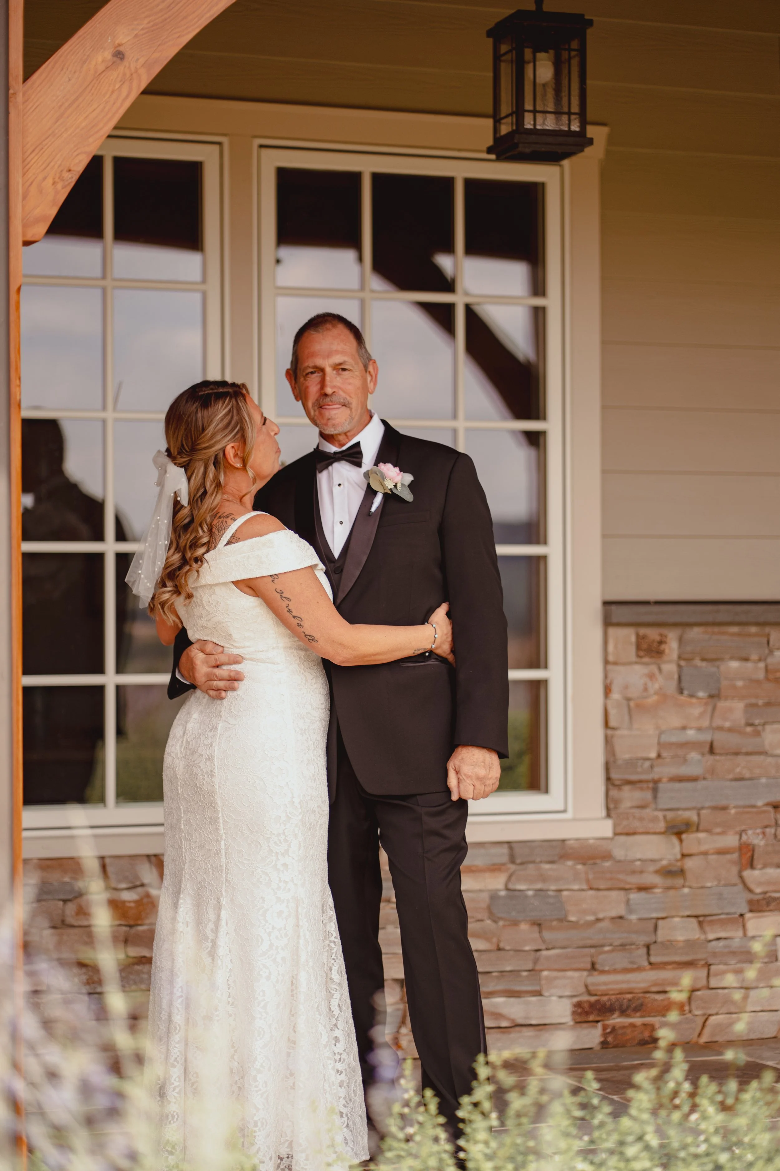 A bride and groom sharing an embrace on their wedding day in front of a large window with a beige and stone exterior wall.