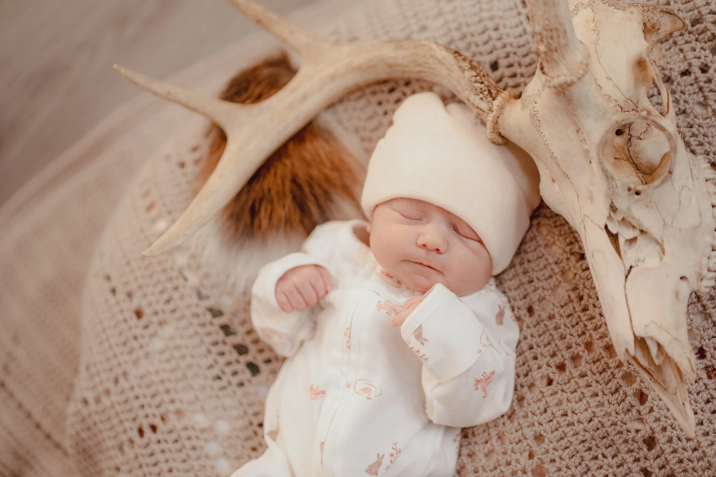 A sleeping baby with a cream hat and pajamas lying on a crocheted blanket, surrounded by antlers and a skull.