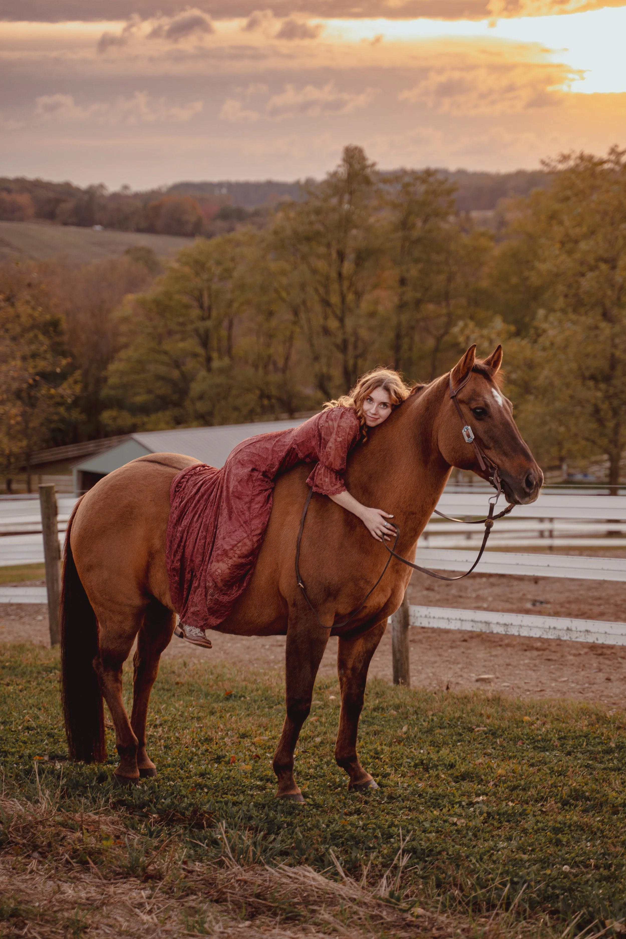 A woman in a long, reddish dress lying on a brown horse, surrounded by a rural landscape with trees and a sunset sky.