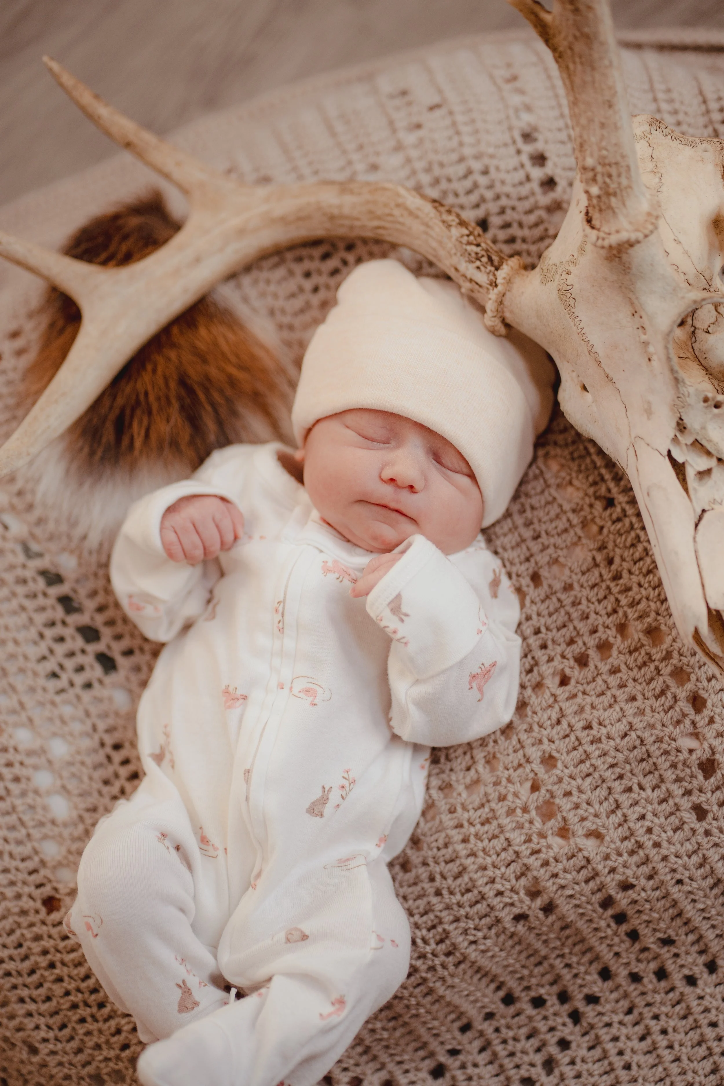 A sleeping newborn baby dressed in a cream-colored onesie with small animal prints, wearing a white hat, lying on a crocheted blanket surrounded by antlers and a skull.
