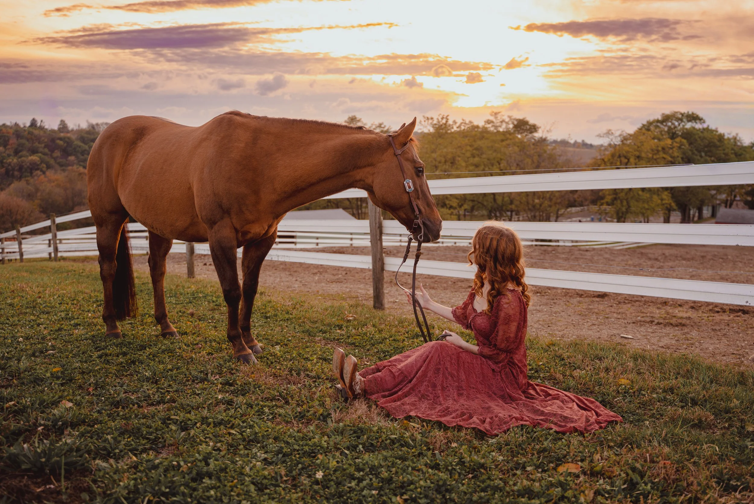 A woman with long red hair wearing a vintage red dress sitting on grass, reaching out to a brown horse, in a rural farm setting at sunset.