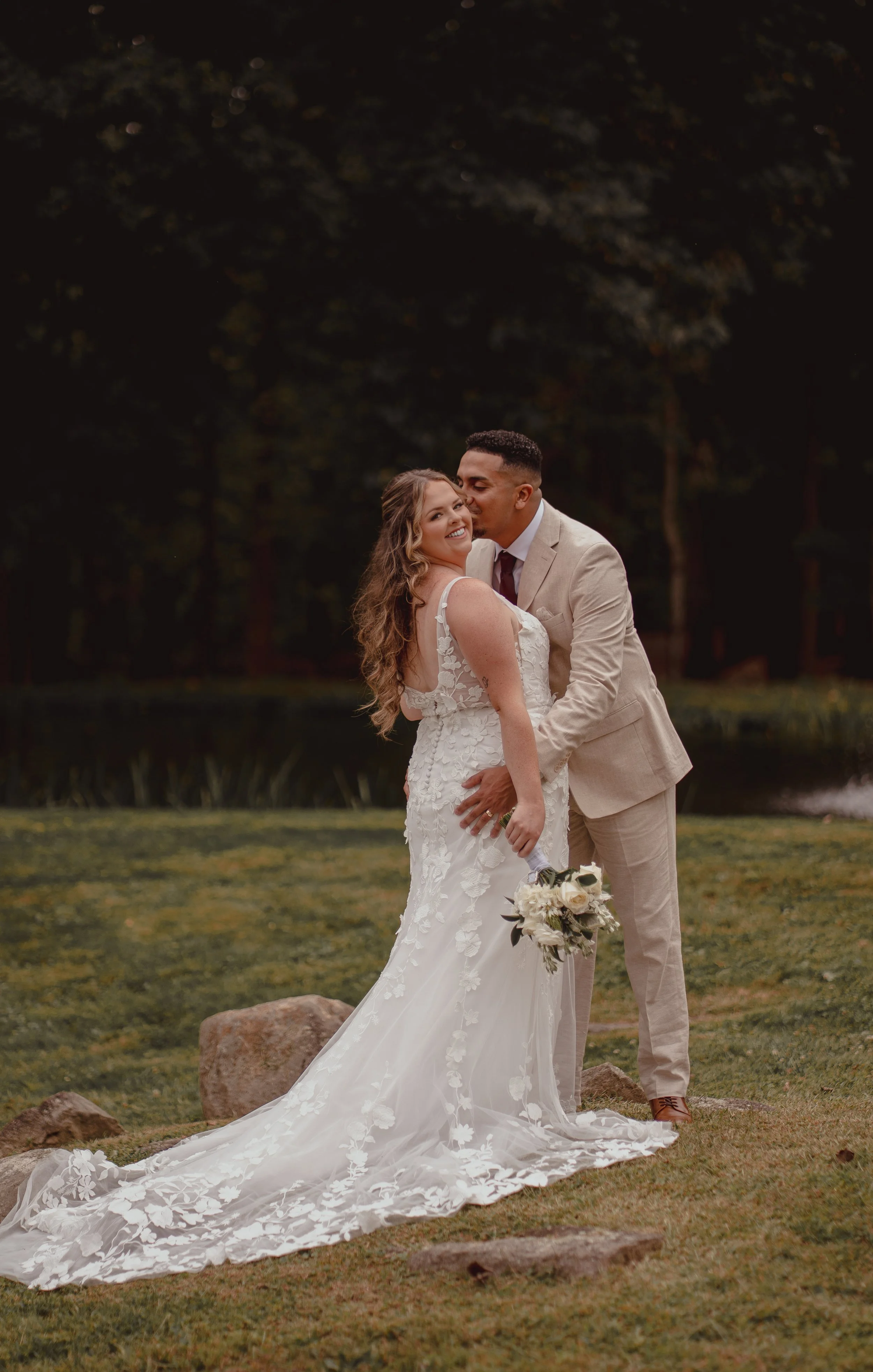 A newlywed couple standing on grass near rocks during sunset. The bride is holding a bouquet and wearing a white lace wedding dress, while the groom is in a beige suit. The groom is kissing the bride on the cheek, and they are smiling happily.